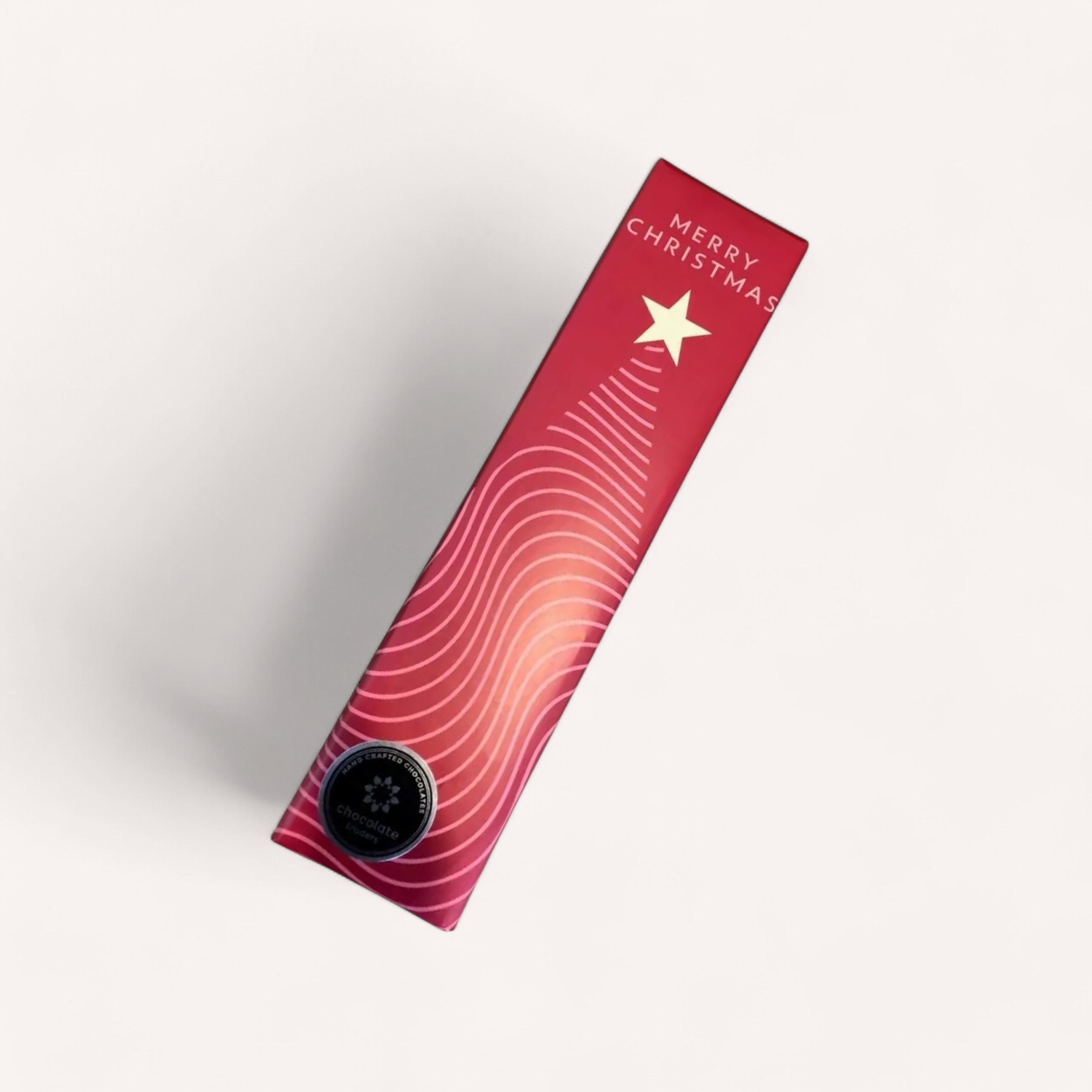 The product, "Merry Christmas Chocolates" by Chocolate Traders, is packaged in a red rectangular gift box. When viewed from the side, the box features a wavy white line that forms a stylized Christmas tree topped with a star. The words "Merry Christmas" are elegantly inscribed in the top right corner. Near the bottom of the design is a small circular emblem adorned with a star, subtly indicating that delectable chocolates await inside. The background is kept plain white to accentuate these designs.