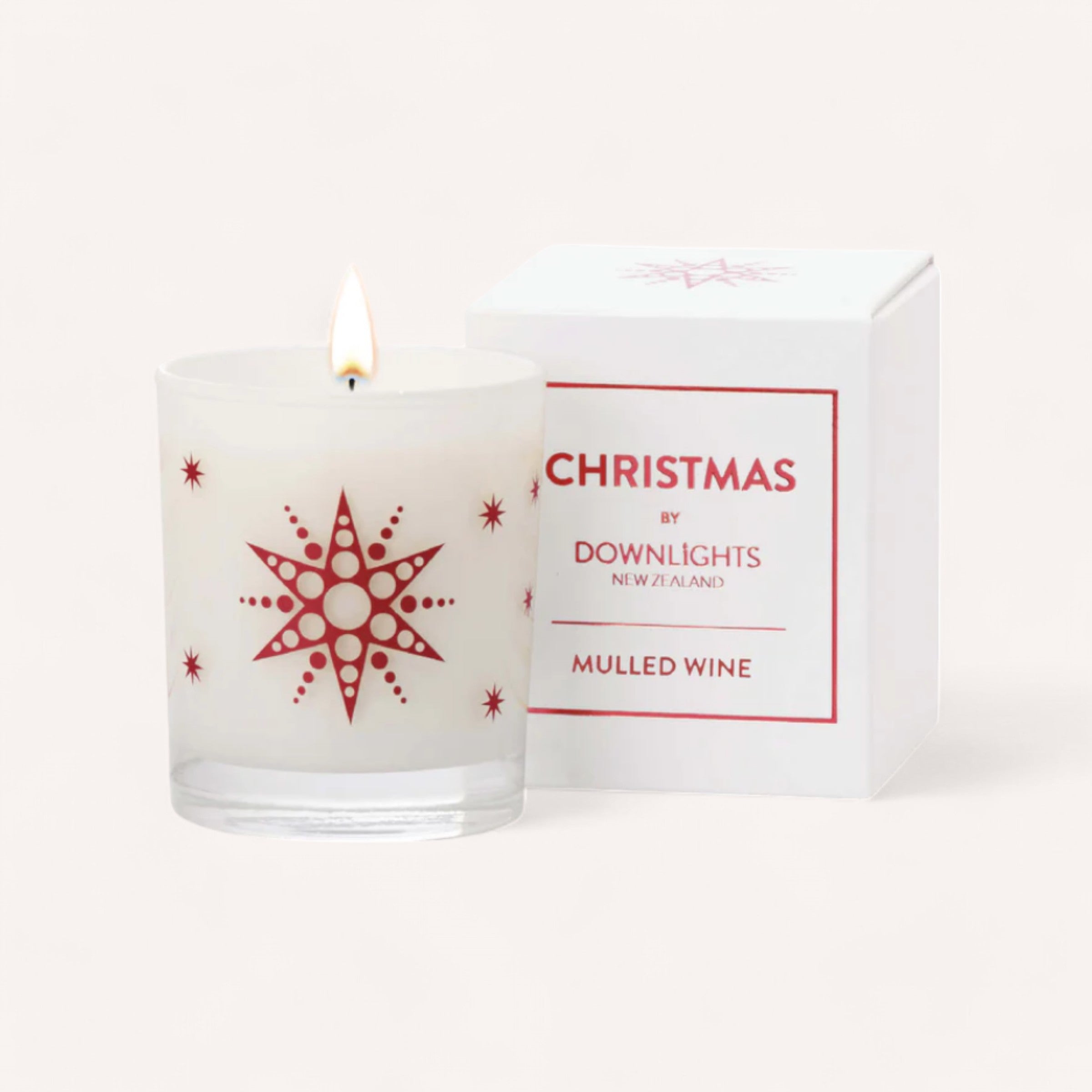 A lit Mulled Wine Christmas Candle by Downlights, enclosed in a glass holder decorated with red stars and snowflake patterns, casts a warm glow. Next to it is a white box labeled "Christmas by Downlights New Zealand, Mulled Wine." The air is filled with the fragrance of orange and clove against the soft cream-colored backdrop.