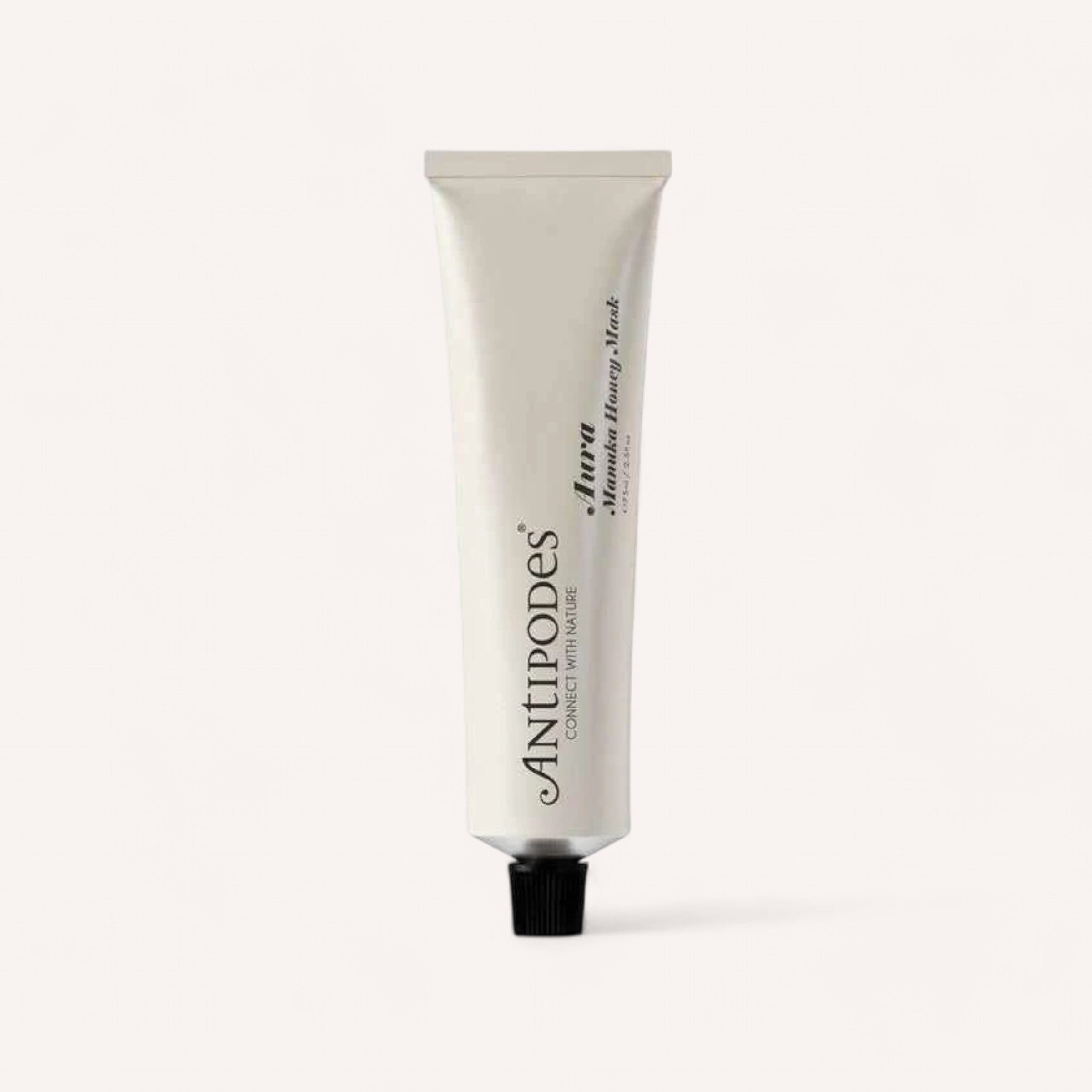 A white tube of Aura Manuka Honey Mask by Antipodes, featuring a black cap, is placed against a simple light backdrop. This face mask, infused with manuka honey, showcases the brand and product information in black text, providing your skin with a hydrating boost.