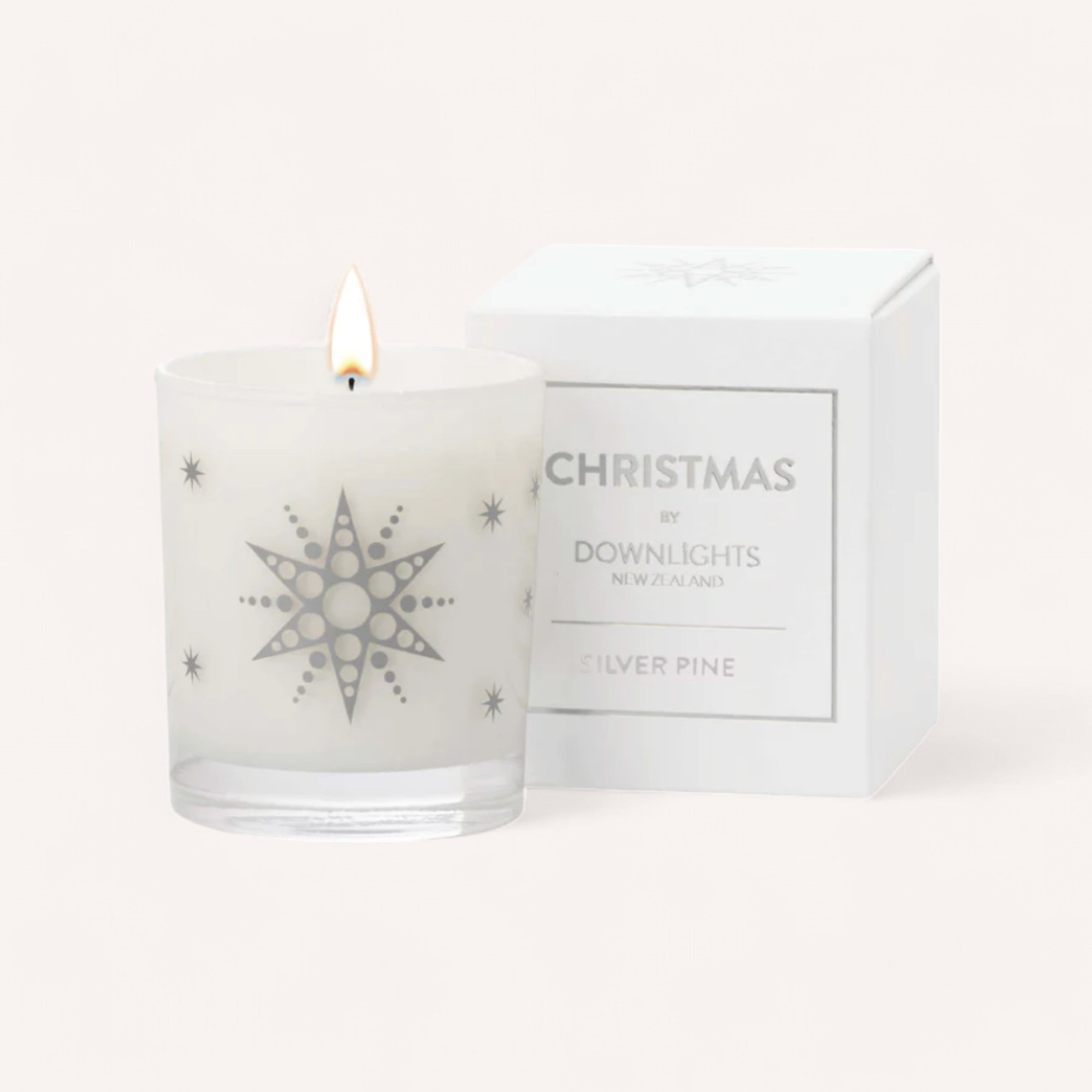 A white candle in a glass holder is alight, showcasing a silver star design with circular accents. It rests next to a matching white box labeled: "Silver Pine Christmas Candle by Downlights," evoking the scent of cedarwood. The background is bright and minimalistic.