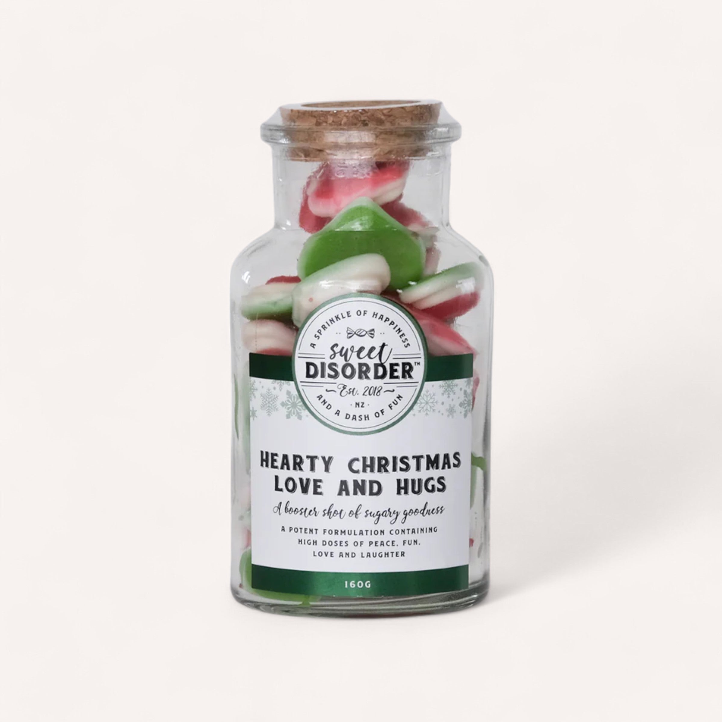 A glass jar with a cork lid holds green and red gummy heart candies. The label displays "Sweet Disorder" with the text "Hearty Christmas Love & Hugs Lollies" below, described as "a booster shot of sugary goodness." The jar rests on a white background with a soft shadow.