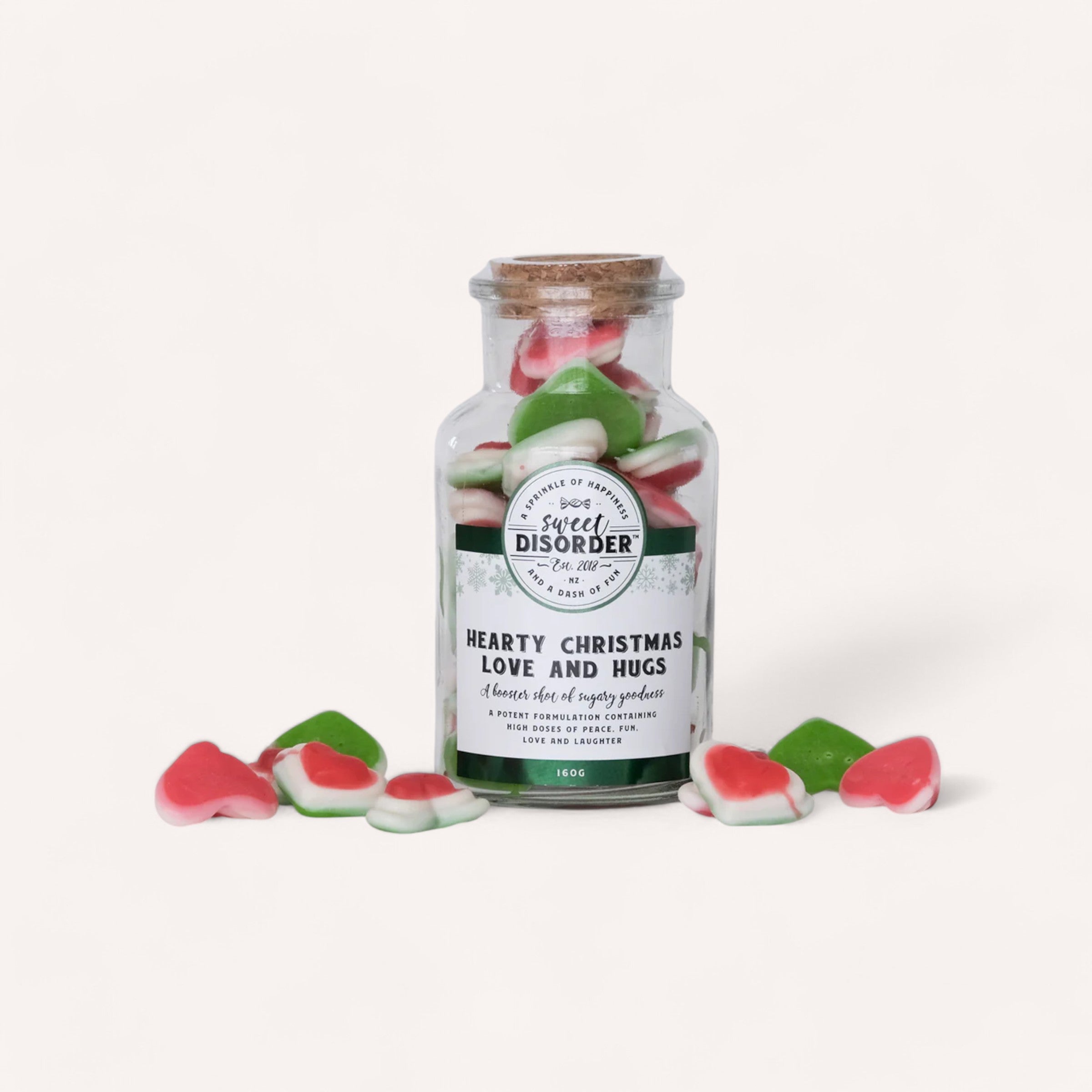 A jar labeled "Sweet Disorder: Hearty Christmas Love & Hugs Lollies" contains red, green, and white gummy heart candies that represent peace. Some candies are scattered around the jar. The label also describes them as "a classic dose of sugary goodness" with a net weight of 140g.