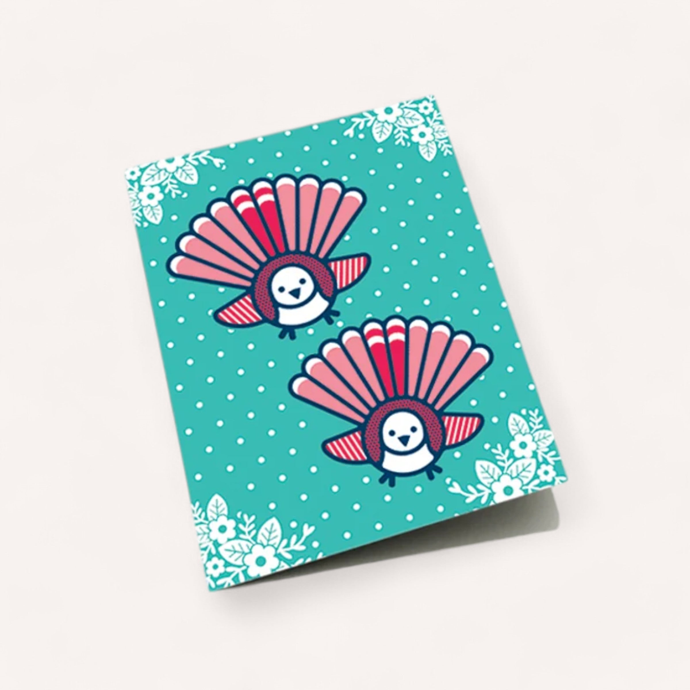 The Fantail Christmas Card by DBL features two stylized peacocks with fan-like pink and red tails set against a teal background. Decorated with white polka dots and floral patterns in the corners, this cheerful and whimsical card comes with a white envelope.
