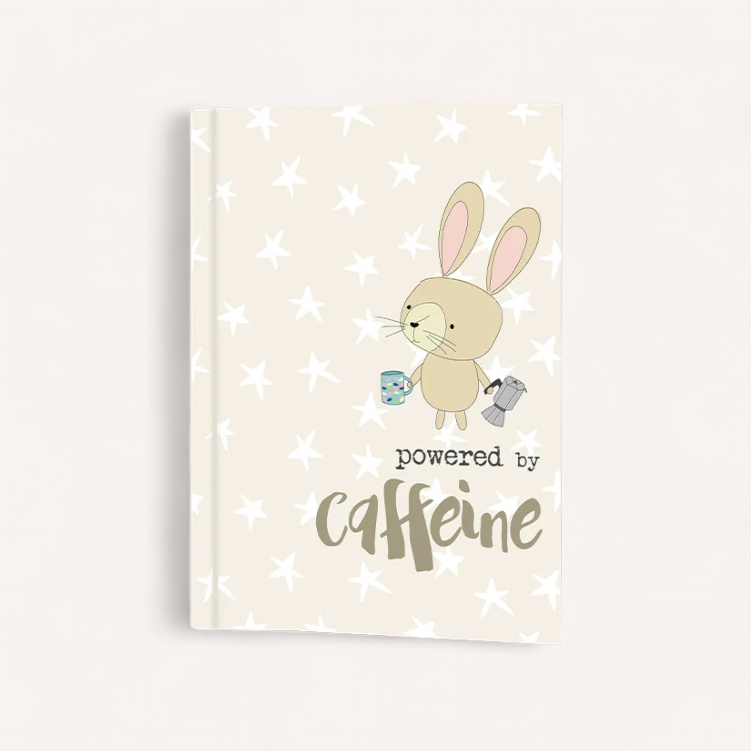 Introducing the Dandelion "Powered by Caffeine Notepad," a delightful A6 notepad featuring a cream cover with white stars. At its heart is an adorable illustration of a bunny holding a blue coffee mug and silver pitcher, highlighted by playful fonts proclaiming it's "powered by caffeine.