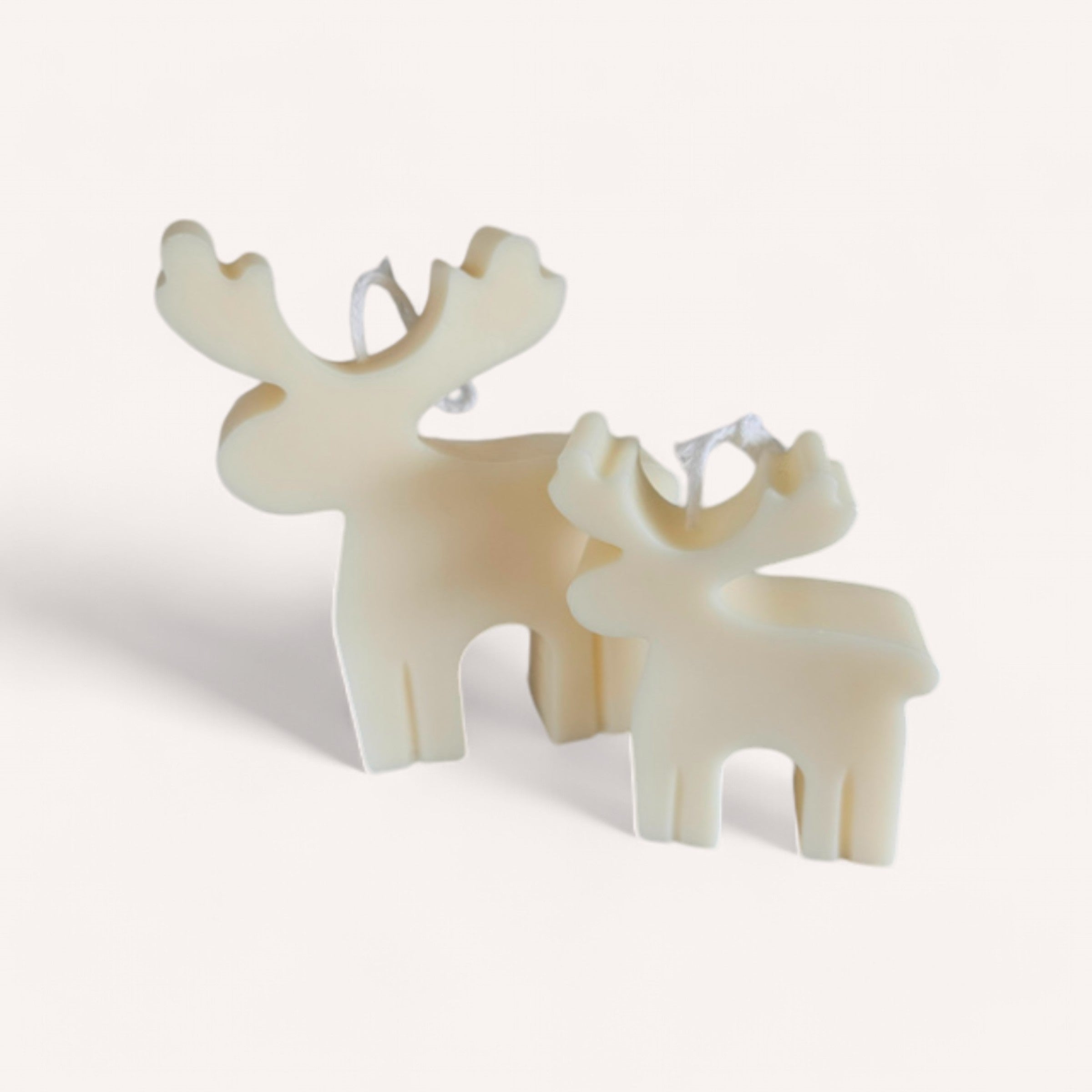 Two cream-colored Wix's Lane Co. Christmas Reindeer Candles are positioned on a white background. The larger candle is slightly behind and next to the smaller one, each showcasing a simple, modern design with visible wicks on top. Shadows cast to the left evoke fresh pine vibes.