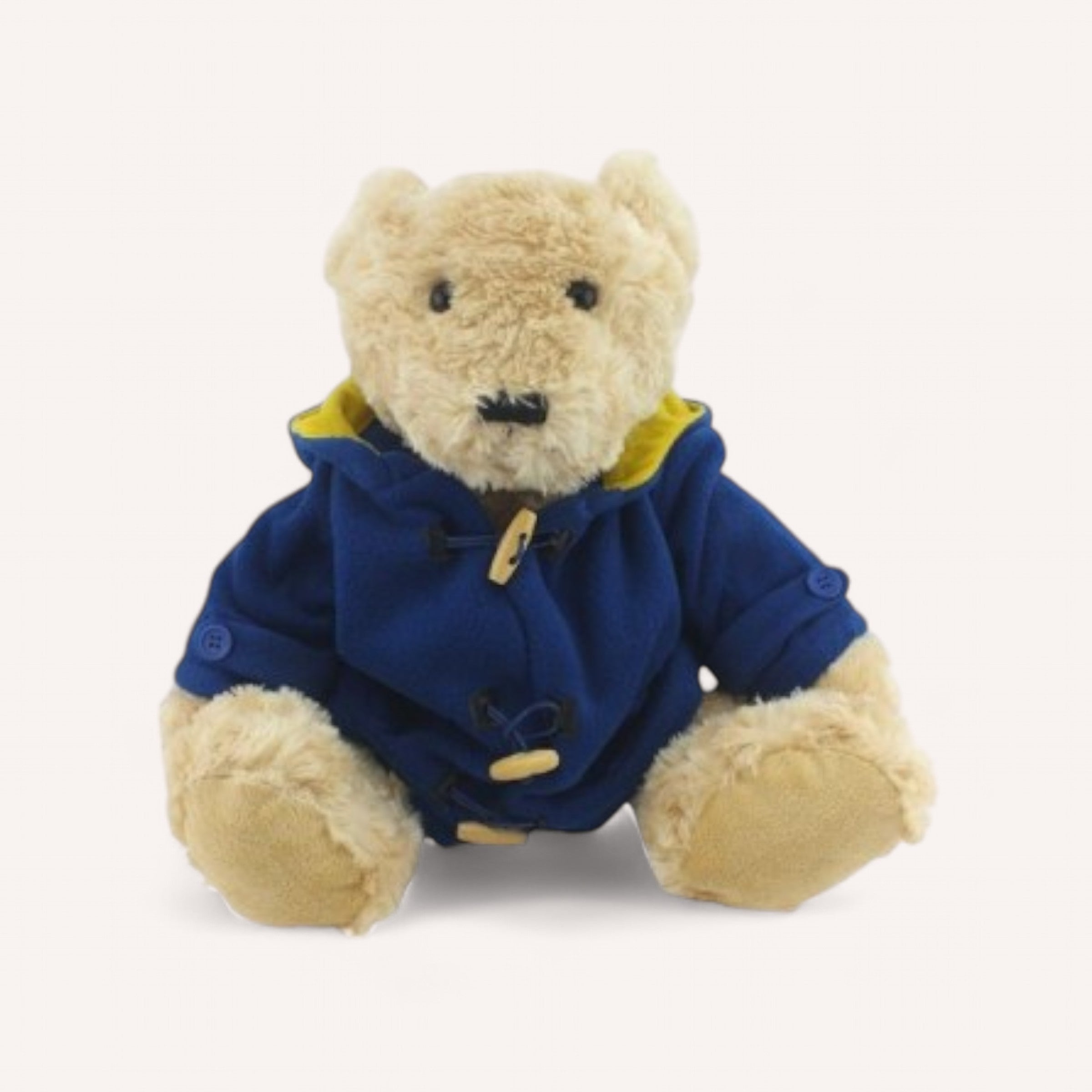 A light brown teddy bear from The Teddy Factory sits upright, wearing the Bear Duffle Coat. This stylish accessory features a blue duffle coat with toggle buttons, a yellow lining, and side buttons on each sleeve. The bear is completed with black stitched eyes and nose, while its paws peek out from under the coat—perfect for chilly days.