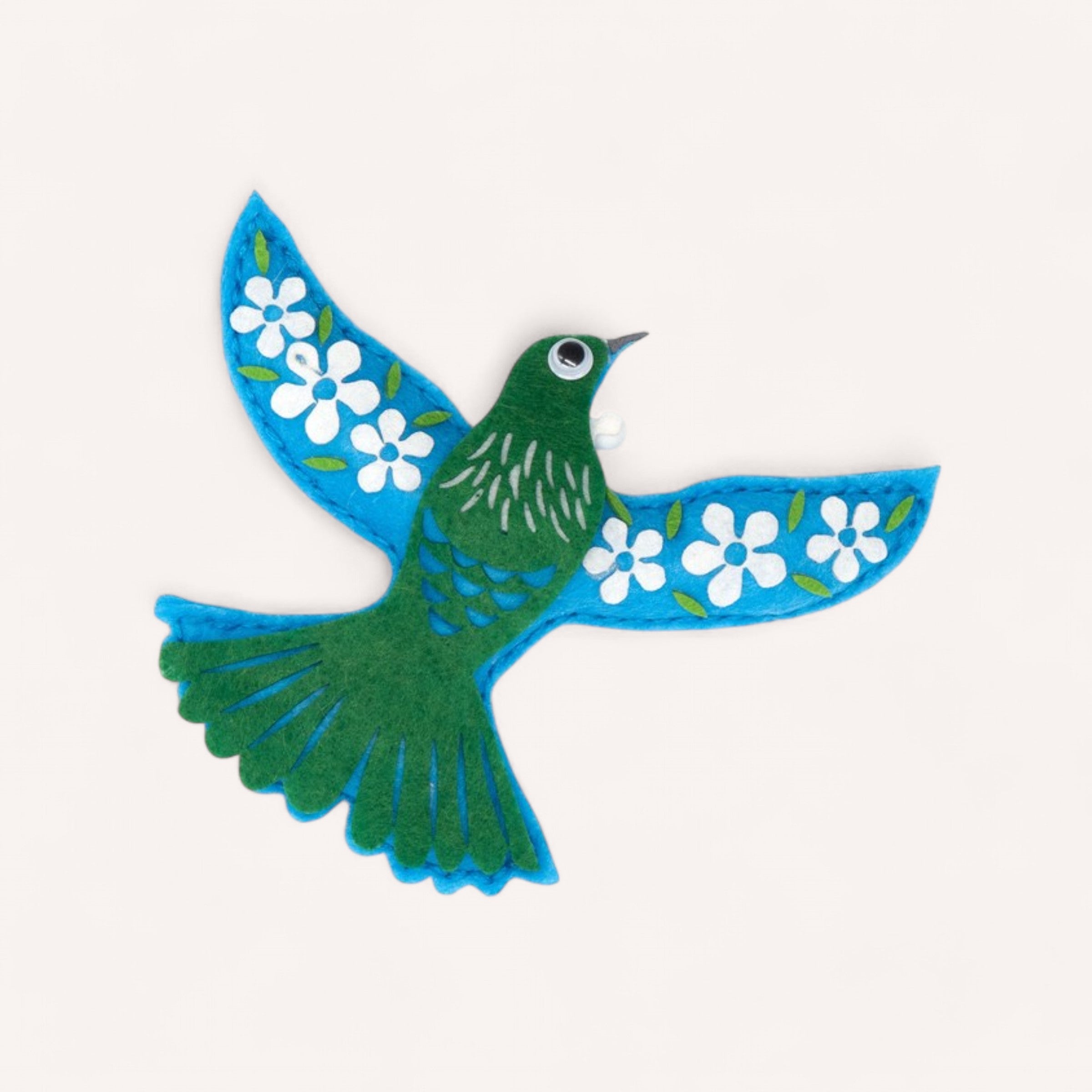 A creative depiction from the Tui Felt Decoration Kit by 100% NZ, featuring a stylized bird with blue wings and a green body. The wings are embellished with white flowers and green leaves, enhancing its artistic flair. The bird's single eye is marked by a white rim and black center, while its tail showcases blue with green accents against a simple white backdrop.