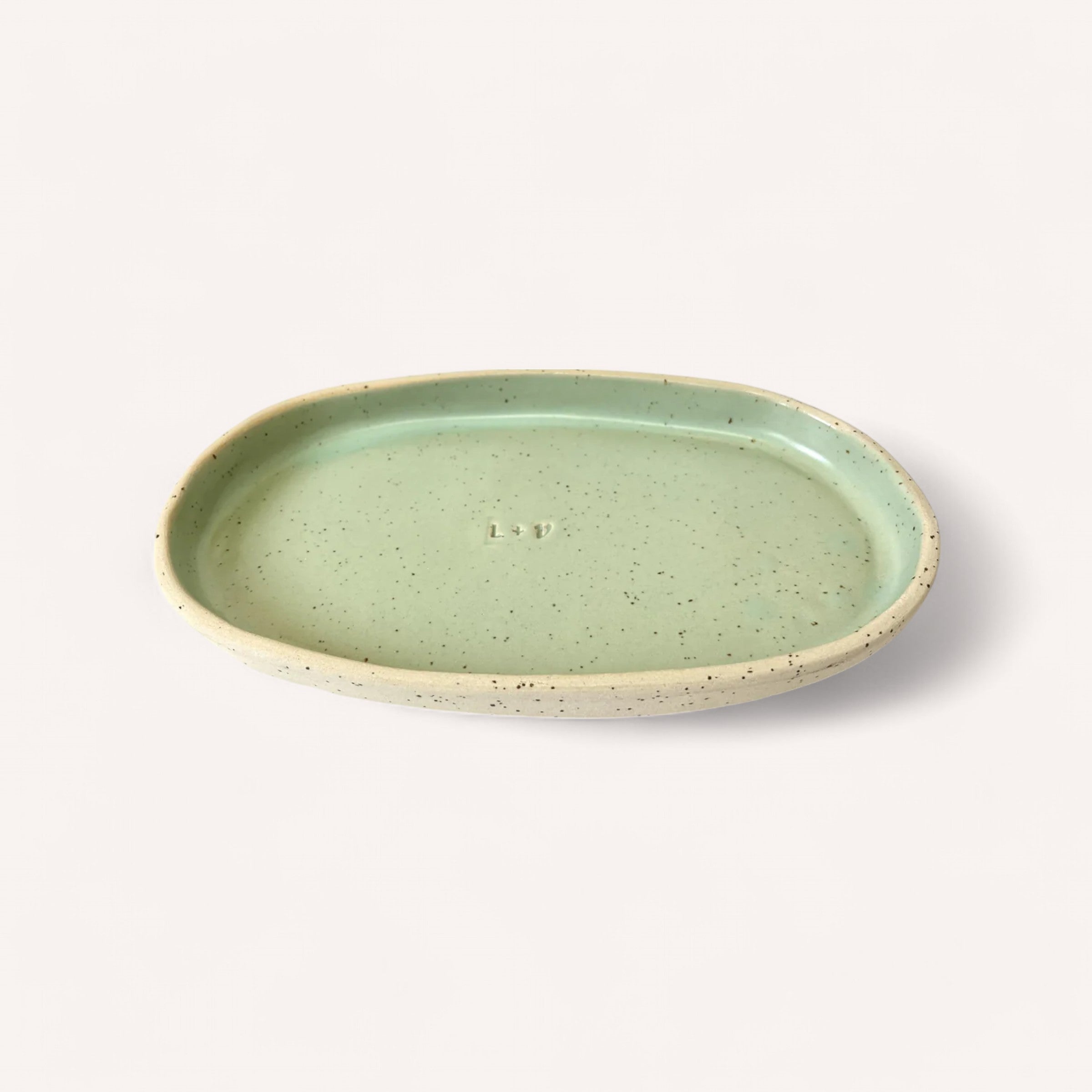 The Handmade Ceramic Tray by Light + Vessel is a speckled, oval-shaped ceramic piece featuring a smooth light green interior and a textured beige rim. The center showcases an embossed logo or initials. Set against a plain, light background, this handcrafted tray highlights its minimalist and artisanal design.