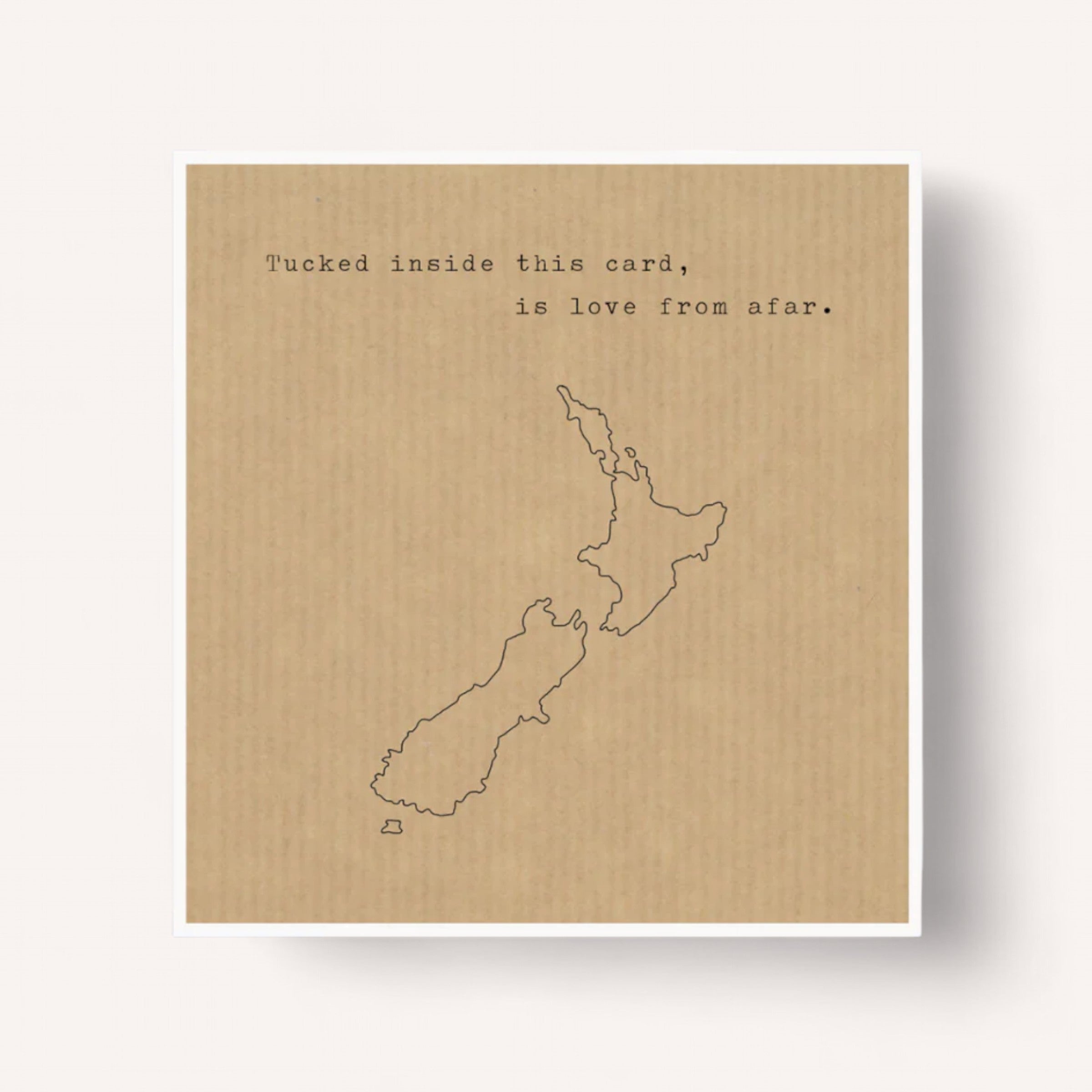 The "Tucked Inside NZ Card" by EmKo showcases a minimalist aesthetic with a beige background and a simple black outline of New Zealand. The text above reads, "Tucked inside this card is love from afar." Accompanied by a white envelope, it offers a heartfelt design and is blank inside for your personal message.