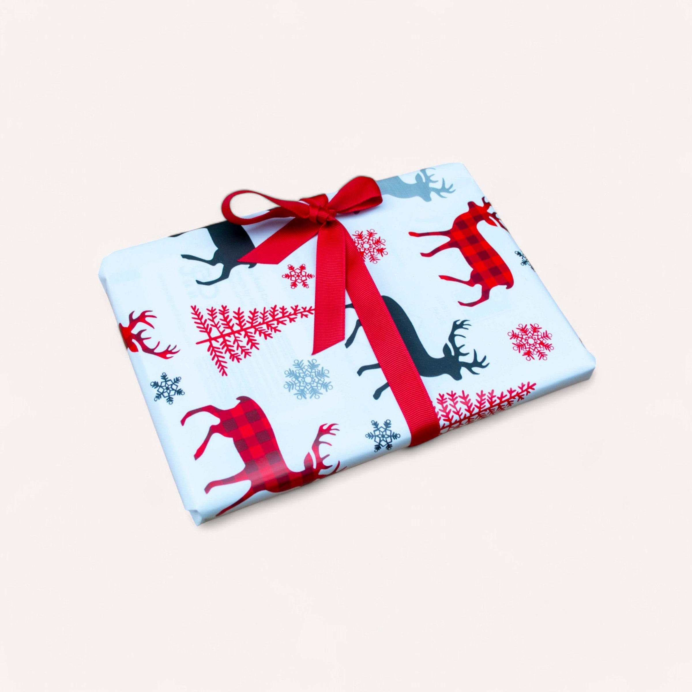 A rectangular gift, expertly wrapped with Gift Wrapping from giftbox co., features premium white paper embellished with red and black reindeer and snowflake patterns. It is tied with a red ribbon that forms a bow on top, presenting a refined look against a plain white background while highlighting the use of eco-friendly recycled materials.