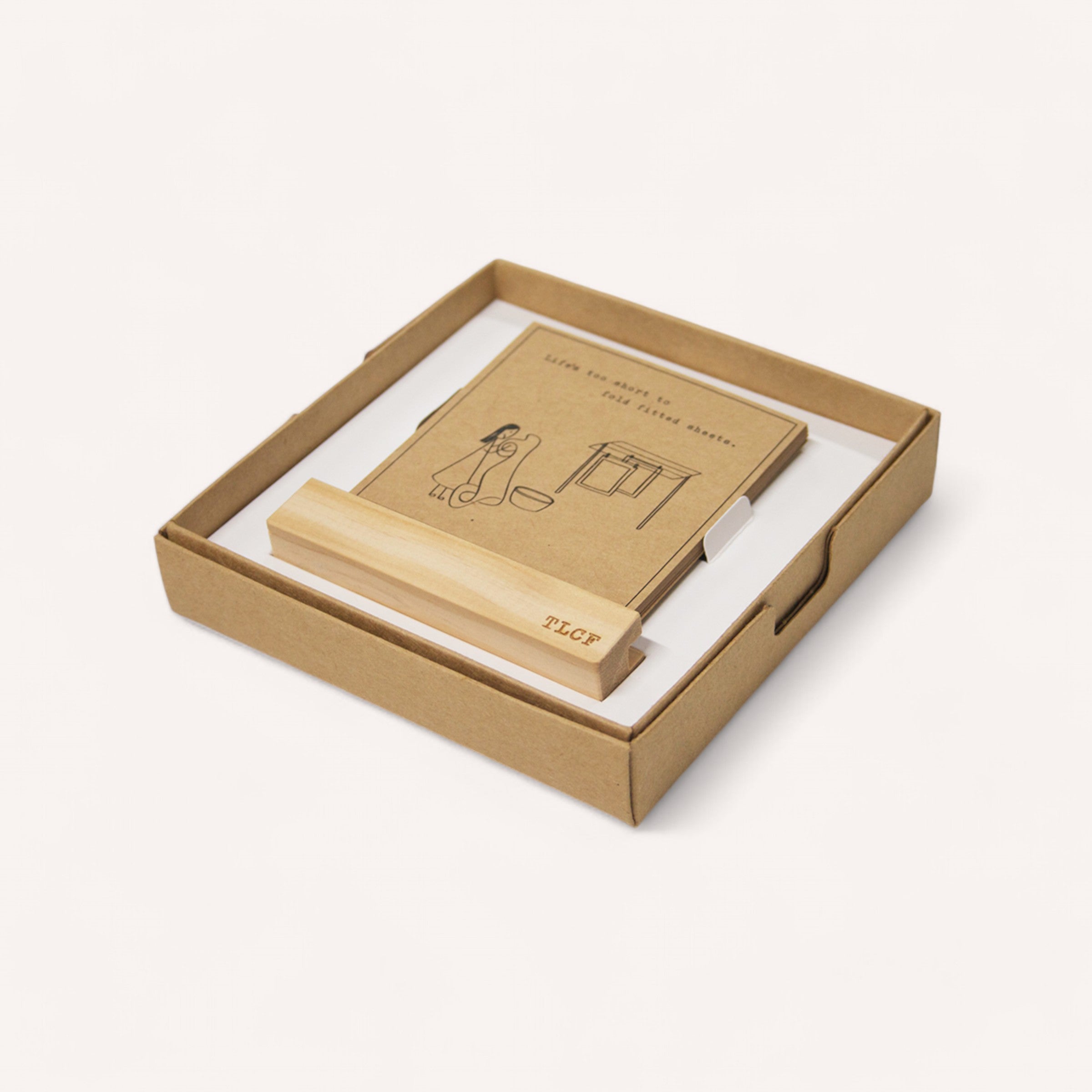 An off-white cardboard box from EmKo features the "Little Squares of Positivity Pack," which includes a wooden stand engraved with "TLFCF" and a book adorned with a cover illustration of someone cleaning. The text on the cover reads, "Life isn't just about the big clean sweep.