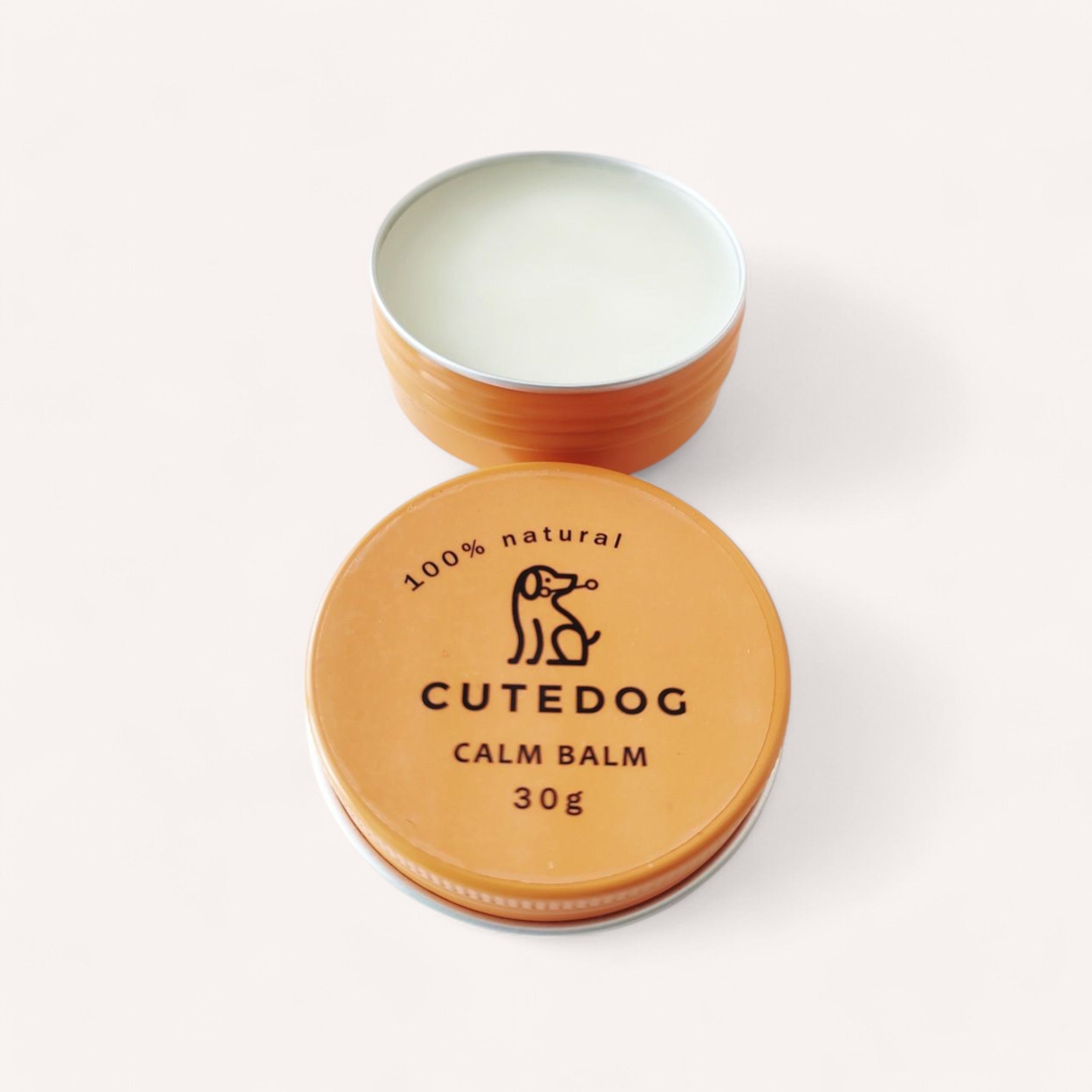 The image features an open tin of "Dog Balm by Cute Dog." The lid, positioned next to the container, is orange with a minimalist black dog logo and text stating "100% natural" and "30g." The balm itself is a smooth, light cream color displayed against a plain white background.