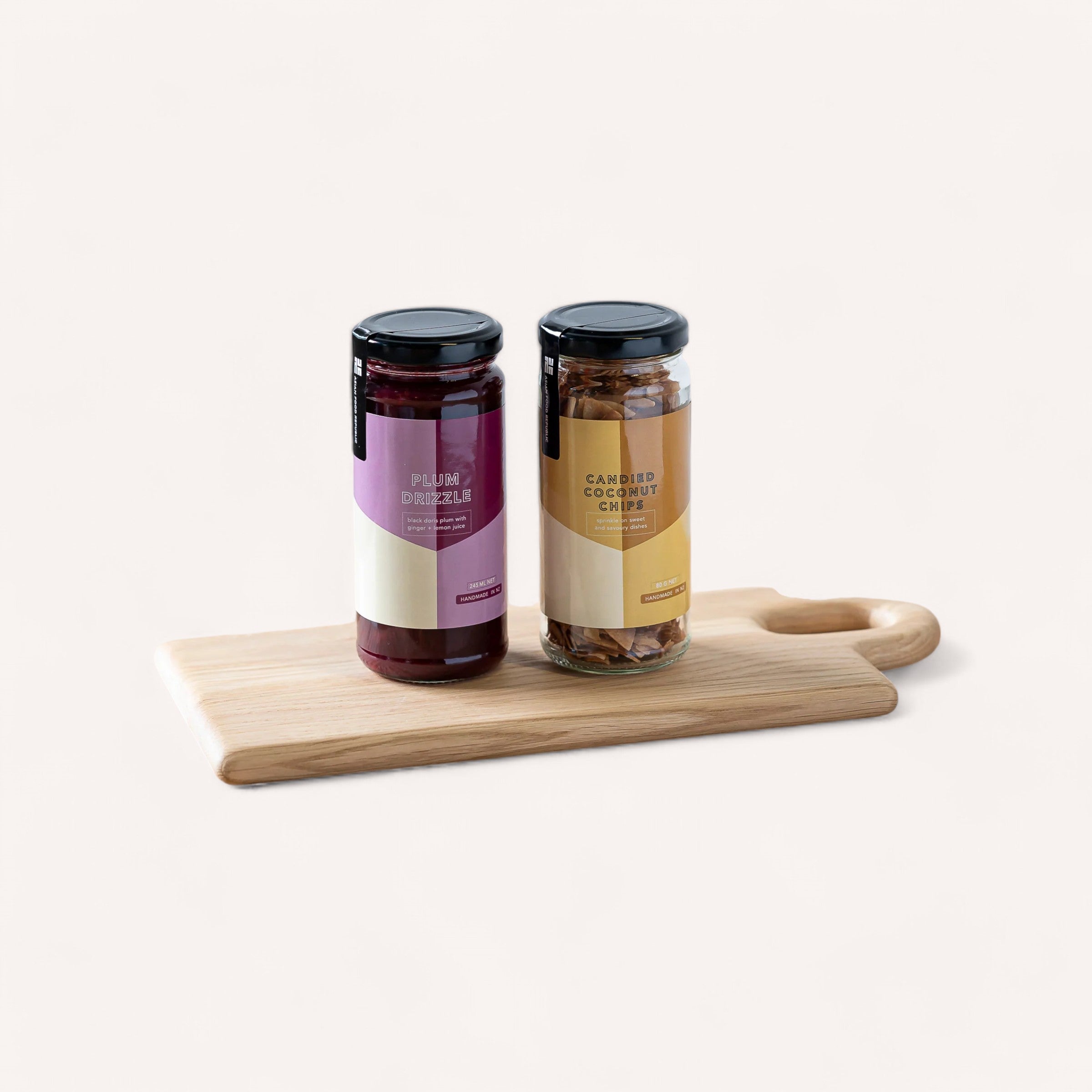 The Christmas Sweet Set from Asian Food Republic makes the perfect holiday gift with its elegant presentation of two jars on a wooden cutting board against a neutral backdrop. The jar on the left, labeled "Plum Preserve," showcases a purple label and holds a rich dark red spread, while the jar on the right, featuring "Candied Chestnut Cream," displays an orange label with a creamy light brown spread. Both jars are topped with sleek black lids.