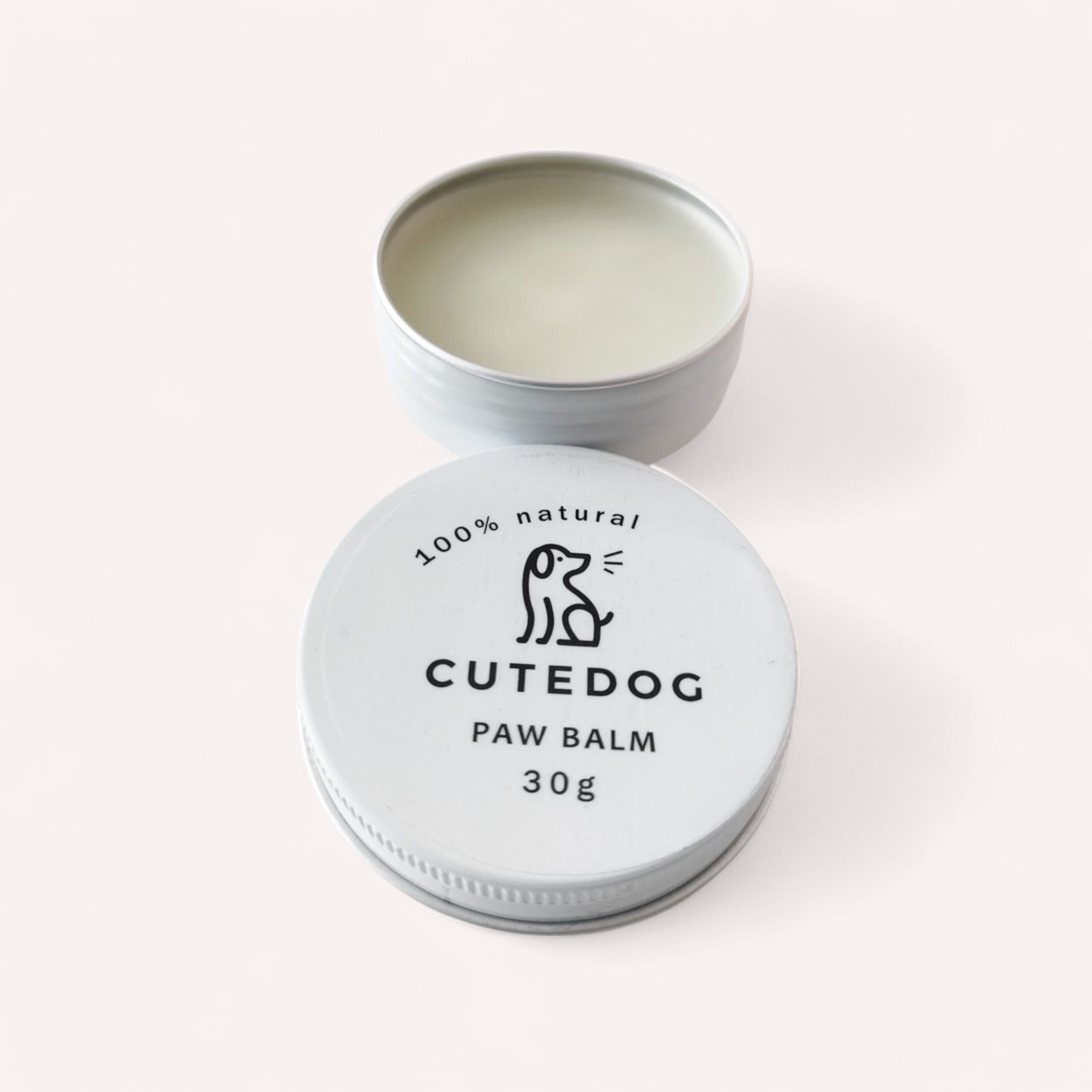 An open small tin of "Dog Balm by Cute Dog" reveals a smooth, creamy balm. The white lid displays a simple line drawing of a dog alongside the text "100% natural" and "30g." The tin is set against a plain, light-colored background.