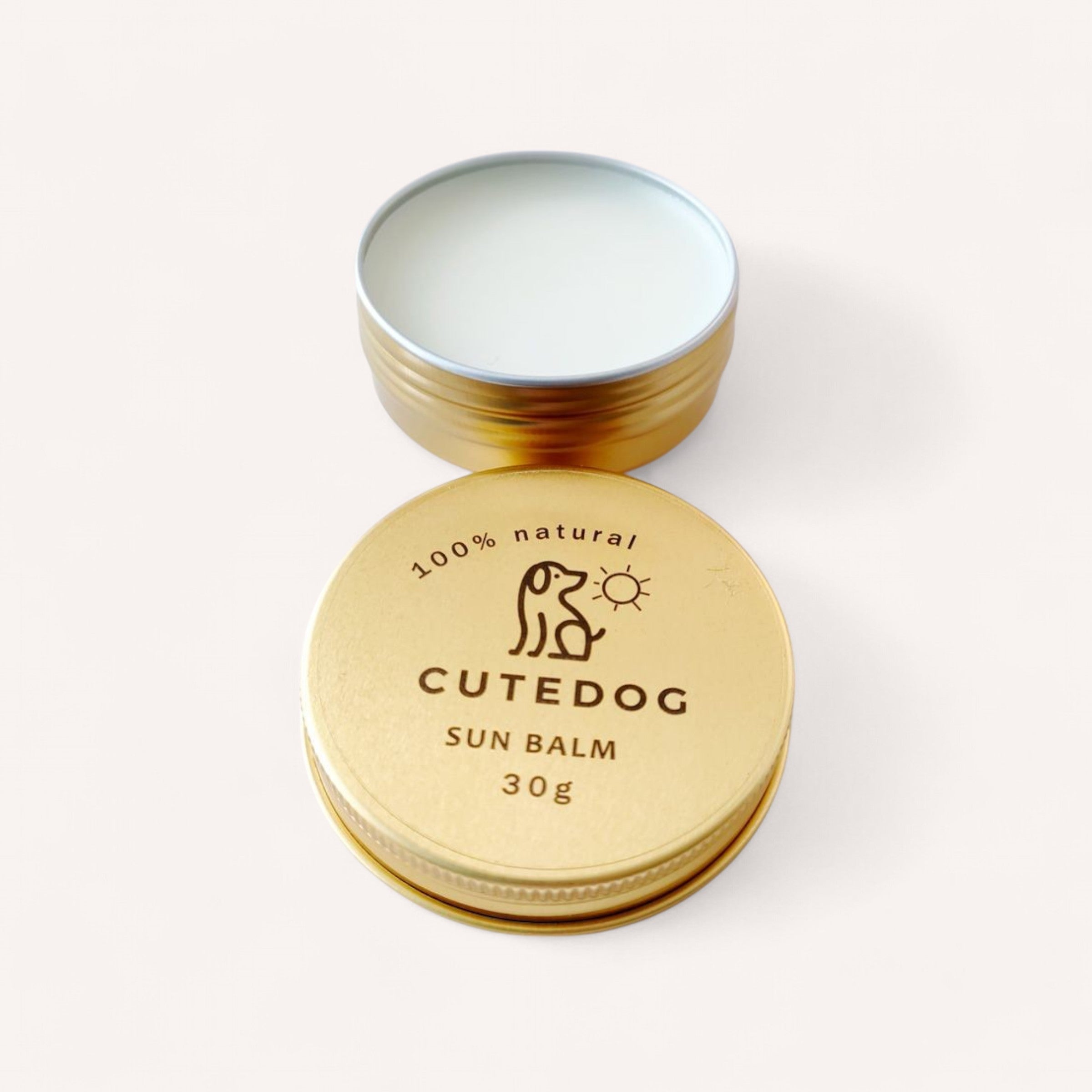 A round, open metal tin labeled "Dog Balm by Cute Dog 30g" reveals a creamy balm inside. The lid, placed beside the tin, features a simple illustration of a dog and sun along with the text "100% natural." The packaging is gold-colored and displayed against a plain white background.