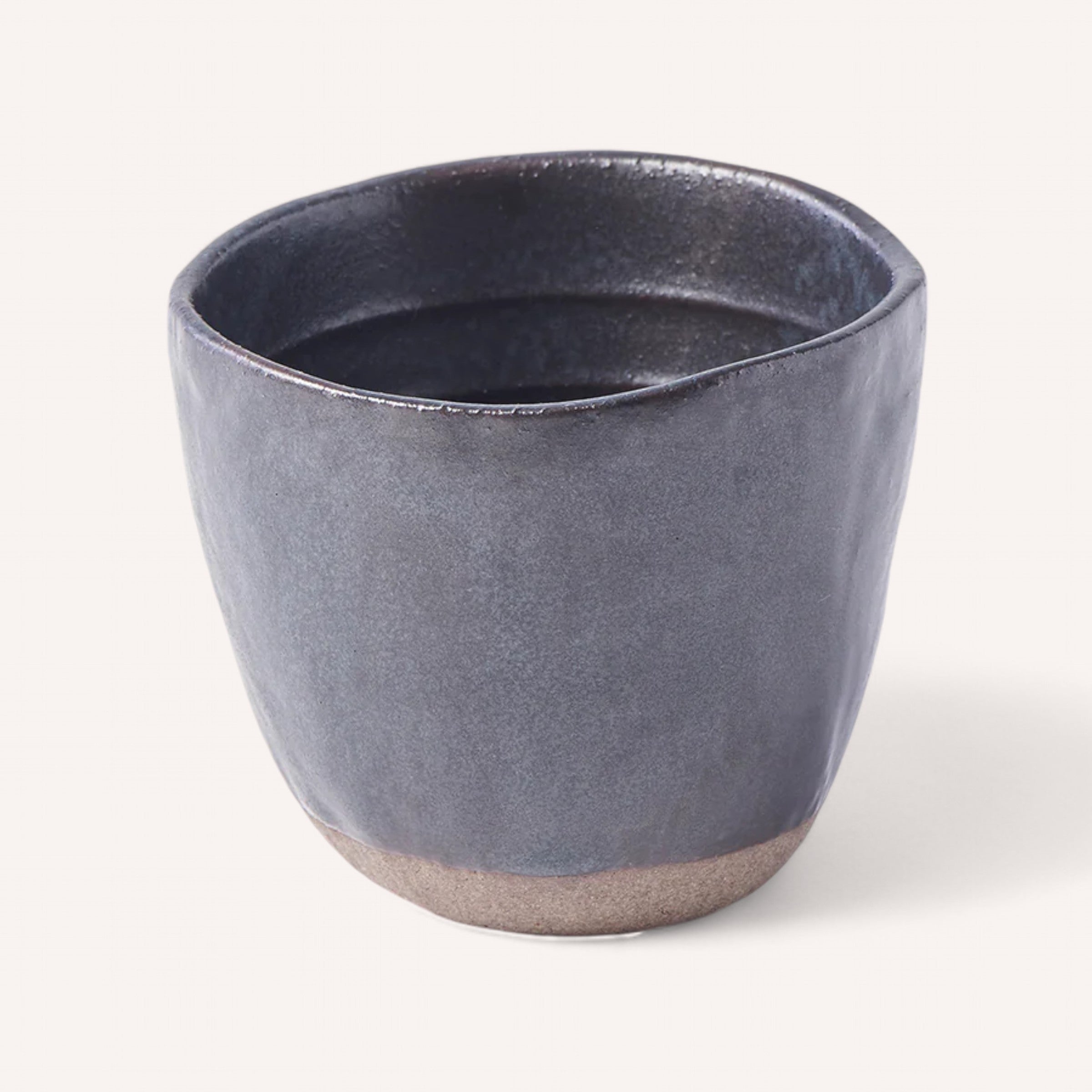 The Naibu Handmade Ceramic Cup features a round design with a rough texture and a dark blue glaze. It has a slightly uneven rim, adding to its rustic charm. The hand-finished surface boasts a subtle sheen and earthy tones at the unglazed base, enhancing its handcrafted appeal against a plain white background.