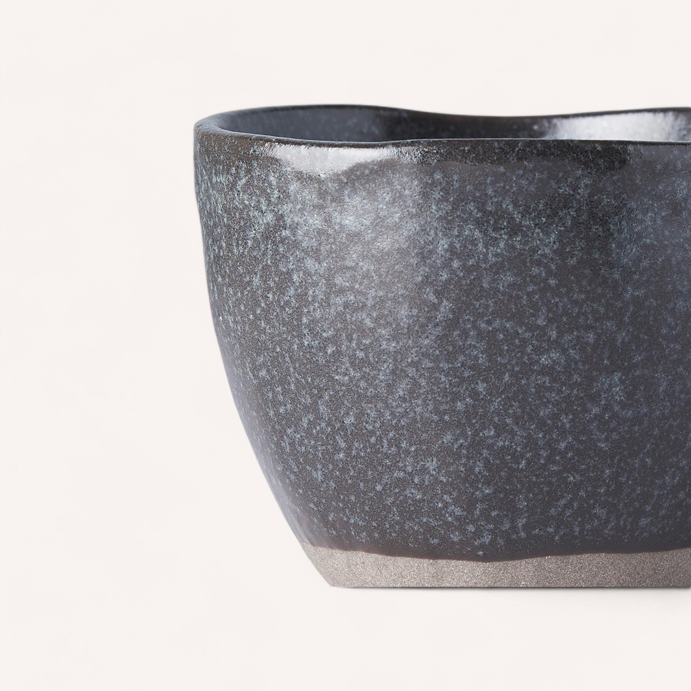 Close-up of a textured ceramic cup featuring a dark, speckled surface. The Naibu Handmade Ceramic Cup resembles Minoyaki porcelain and boasts an uneven, organic shape with a matte finish, transitioning from deep charcoal to lighter speckles. The soft white background accentuates the cup's artistry and craftsmanship.