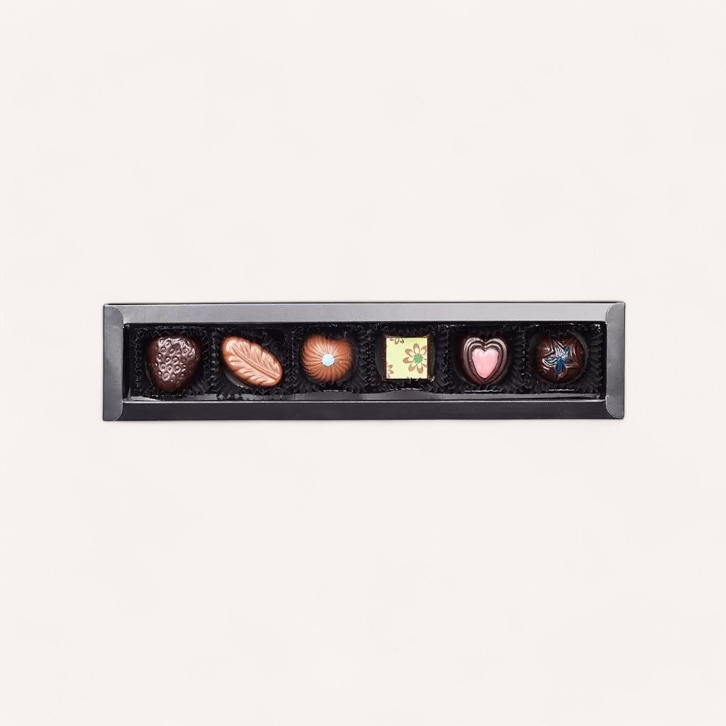 Introducing the Merry Christmas Chocolates by Chocolate Traders: a sleek black rectangular gift box showcasing six distinctive gourmet chocolates. From left to right, discover a heart-shaped piece, a leaf-shaped delight, a dome-shaped treat, a square chocolate adorned with floral patterns, another heart-shaped confection with a pink hue, and finally, a round delicacy featuring a blue swirl on top—all elegantly set against a pristine white background.