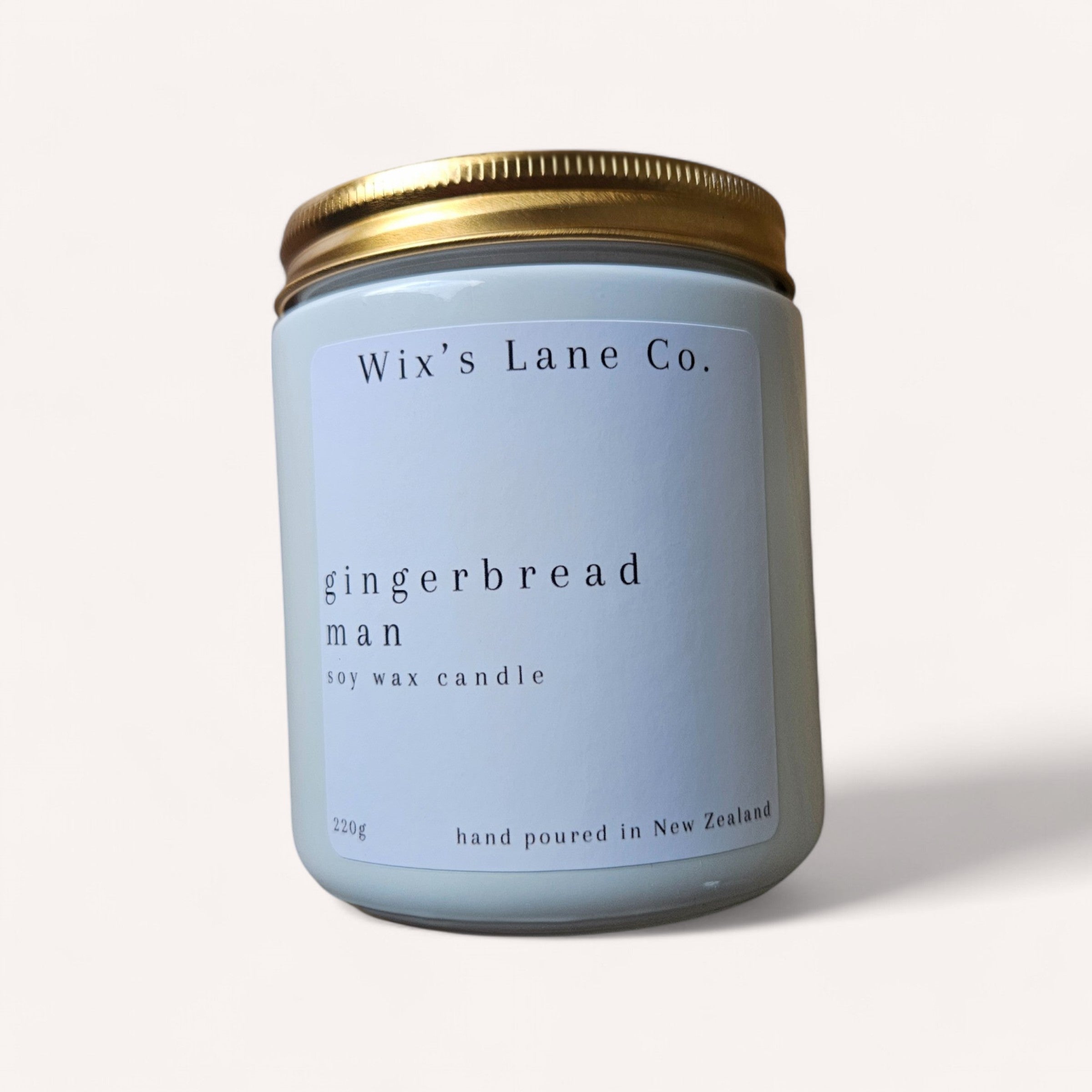 A light blue candle jar with a gold metallic lid, labeled "Christmas Candle by Wix's Lane Co." The text notes it is hand poured in New Zealand and the candle weighs 220g. The background is white.