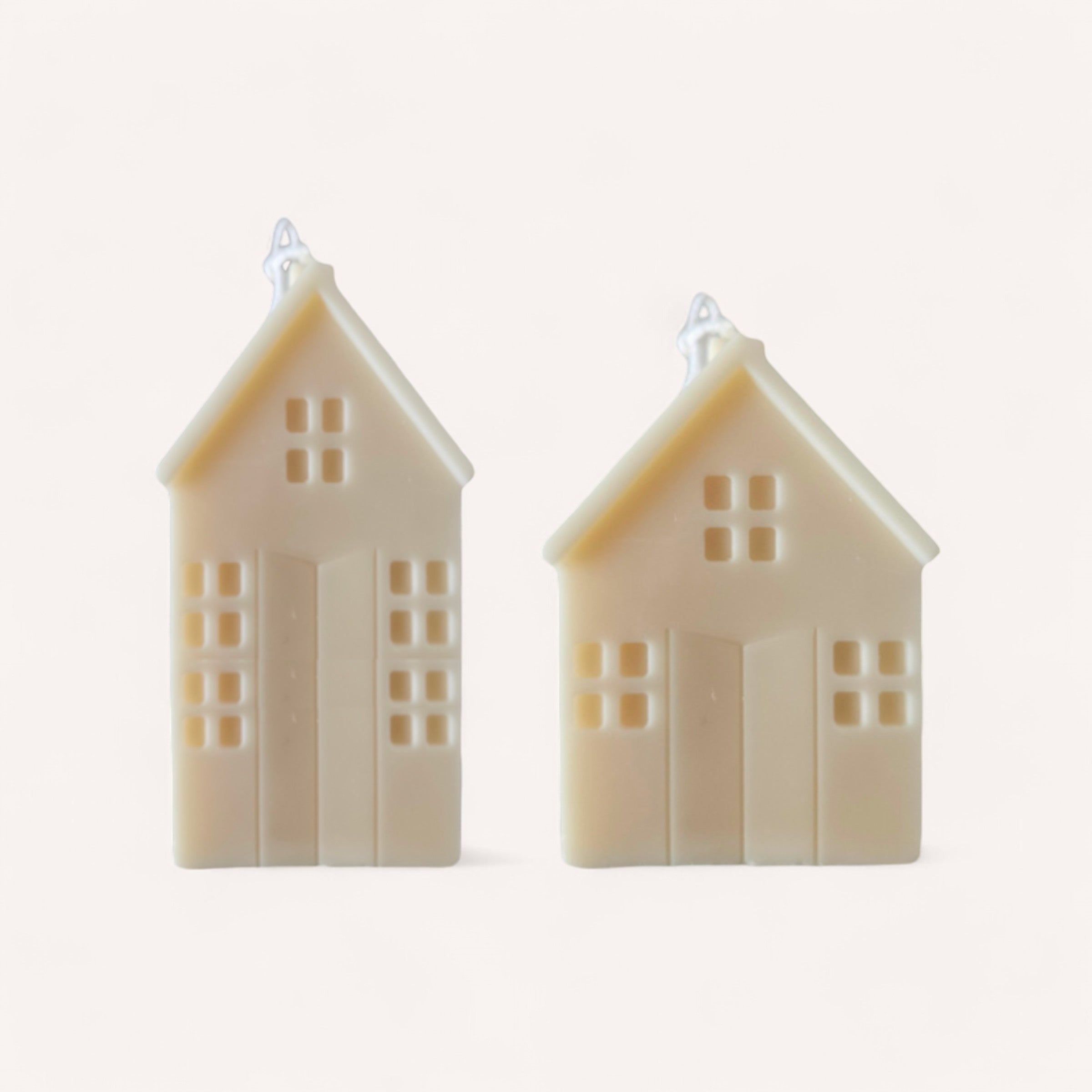 Two creamy beige Christmas House Candles, handcrafted by Wix's Lane Co., are shaped like quaint houses with windows and a door. Each candle features a pitched roof adorned with a small star. The cozy scene is complemented by an off-white background, perfect for evoking the charm of a Christmas candle village.