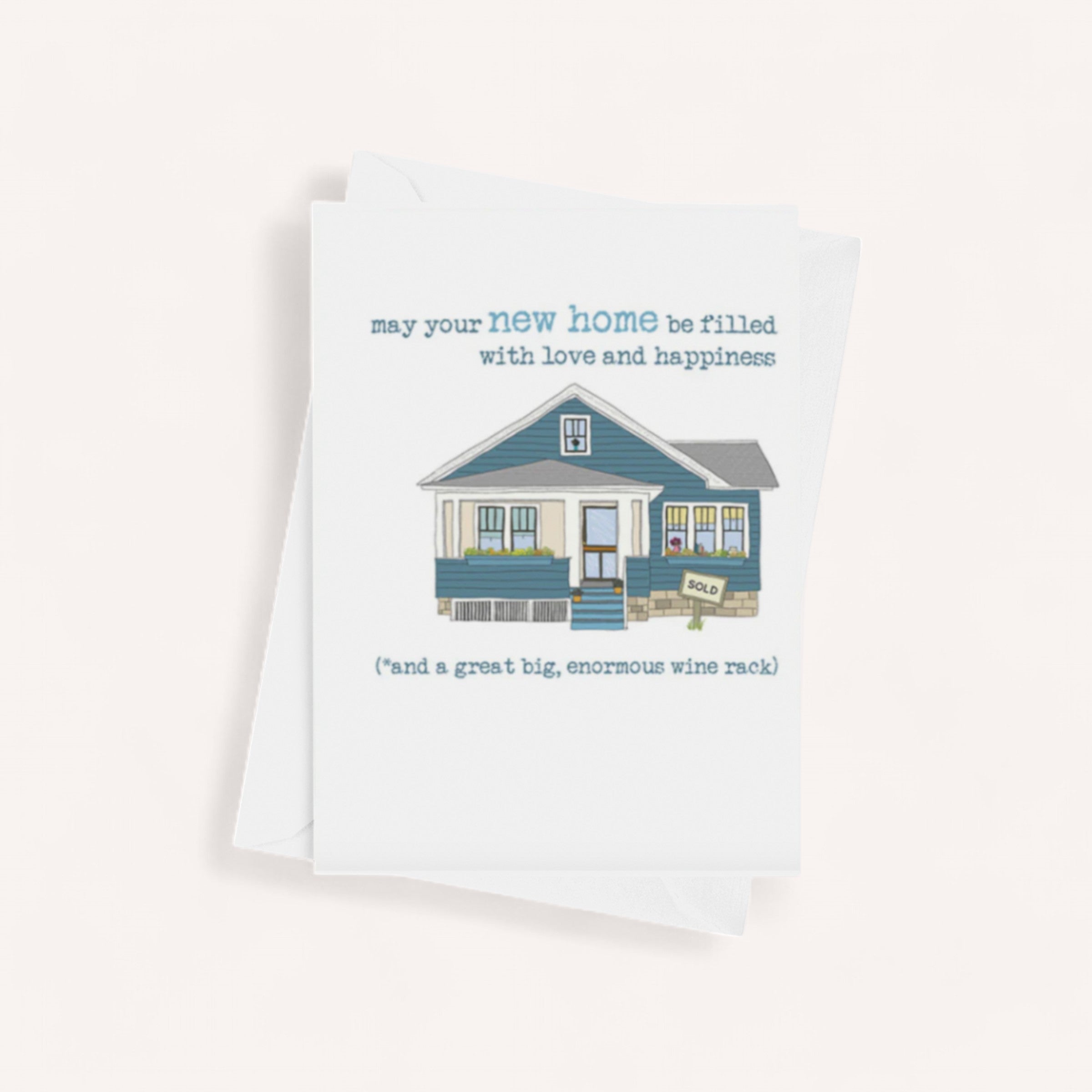 Illustration of a small blue house with a porch and a "SOLD" sign. The text above reads, "may your new home be filled with love and happiness," with a humorous note at the bottom saying, "(*and a great big, enormous wine rack)." The Test by giftbox co. card is placed on top of a white envelope.