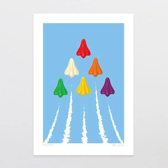 The "Red Leader" framed print by Glenn Jones features seven vibrant paper planes mimicking jet planes in red, orange, yellow, green, blue, purple, and white soaring in a V formation against a blue sky. Each plane is trailed by contrail-like white streams. The image is elegantly presented with a striking white border.