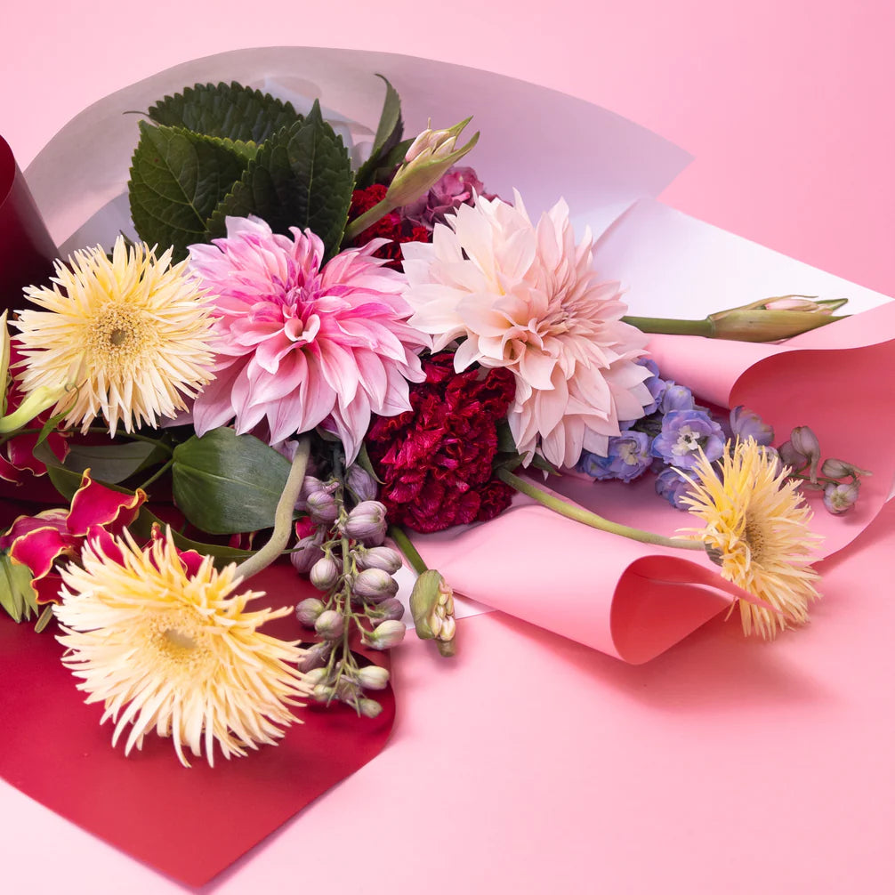 A colorful bouquet of flowers including pink dahlias, yellow spider chrysanthemums, red carnations, blue delphiniums, and green leaves, wrapped in red and white paper with a pink backdrop. This arrangement is part of our bespoke floral collections and includes a handwritten card for a personal touch. Introducing the Florist Choice by Poppy in April.