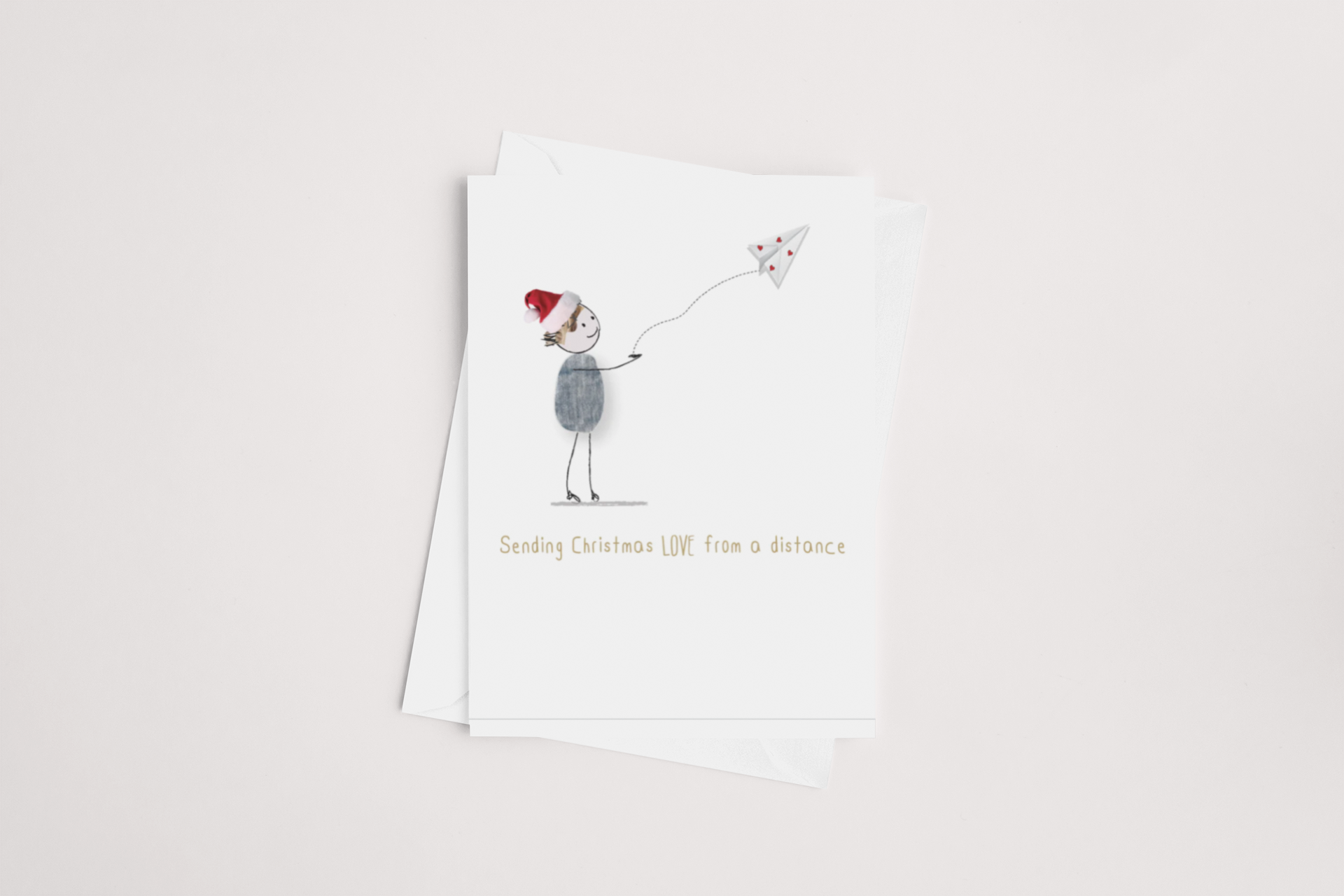 An illustration on the iCandy "Sending Love from a Distance Christmas Card" shows a person in a Santa hat holding a string connected to a paper airplane adorned with a red heart, capturing the essence of "Sending Christmas LOVE from a distance." The card provides ample space for your personalized message with its blank interior against an understated background.