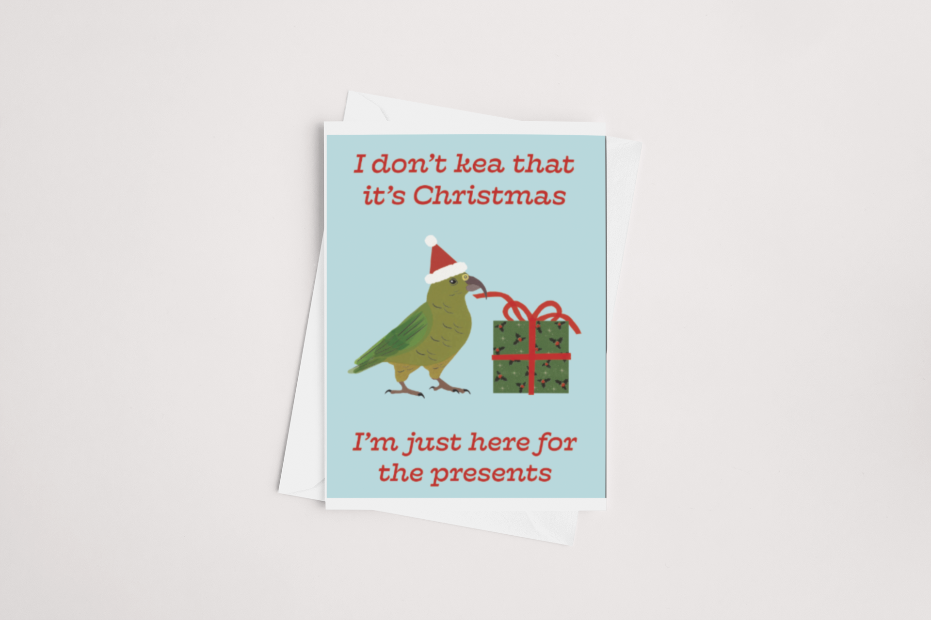 The Kea Christmas Card by Tuesday Print depicts a cartoon kea wearing a Santa hat next to a wrapped gift tied with a bow. The text above reads, "I don't kea that it's Christmas," and below it says, "I'm just here for the presents." The card is blank inside for your personalized message and includes a white envelope.