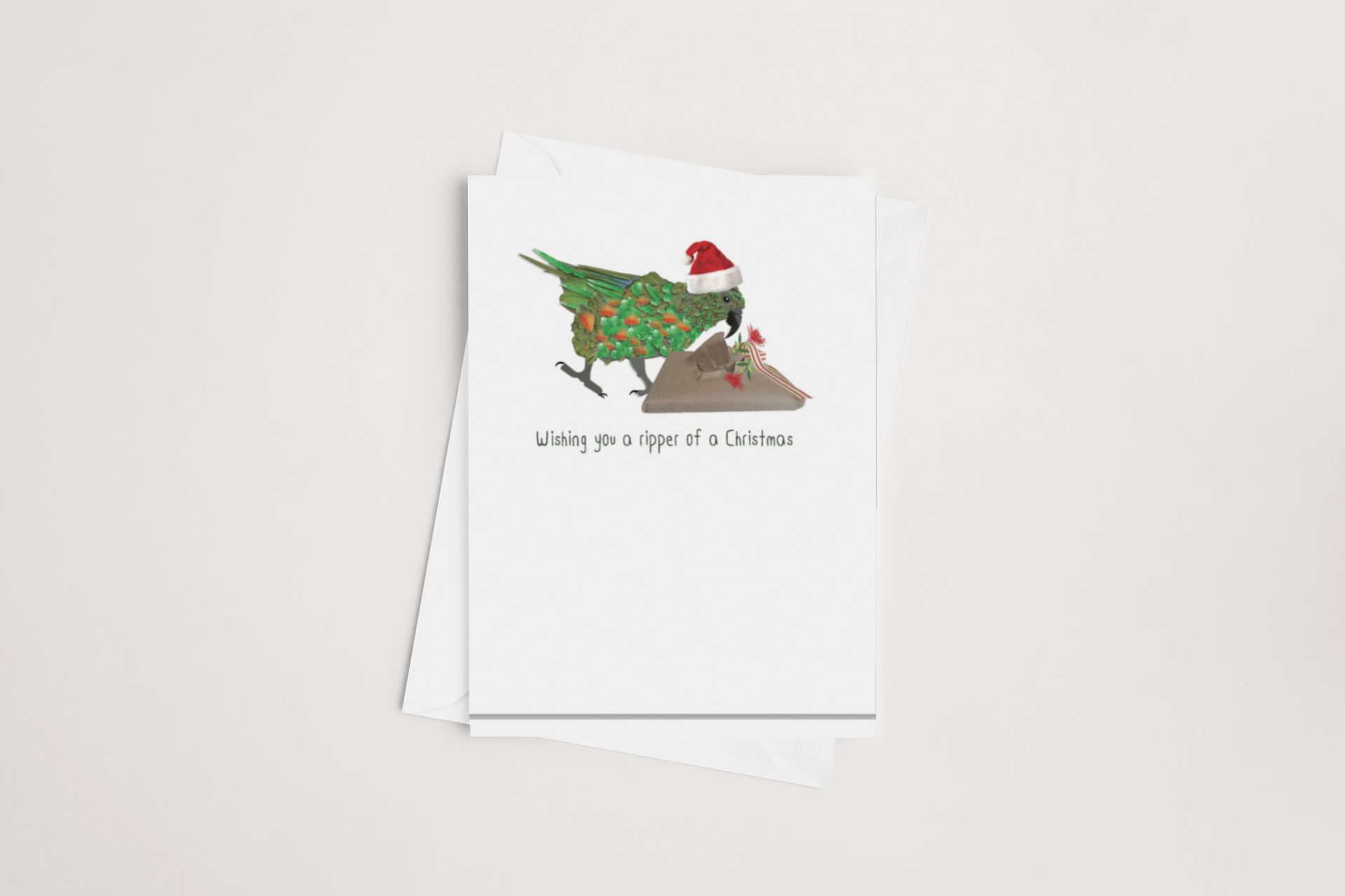 The Ripper Kea Christmas Card by iCandy showcases a cheerful green bird decorated with holly leaves and berries, donning a red Santa hat while holding an envelope. The card's blank interior allows for your personalized message, and the front boldly states "Wishing you a ripper of a Christmas" in black lettering. It comes with a white envelope.