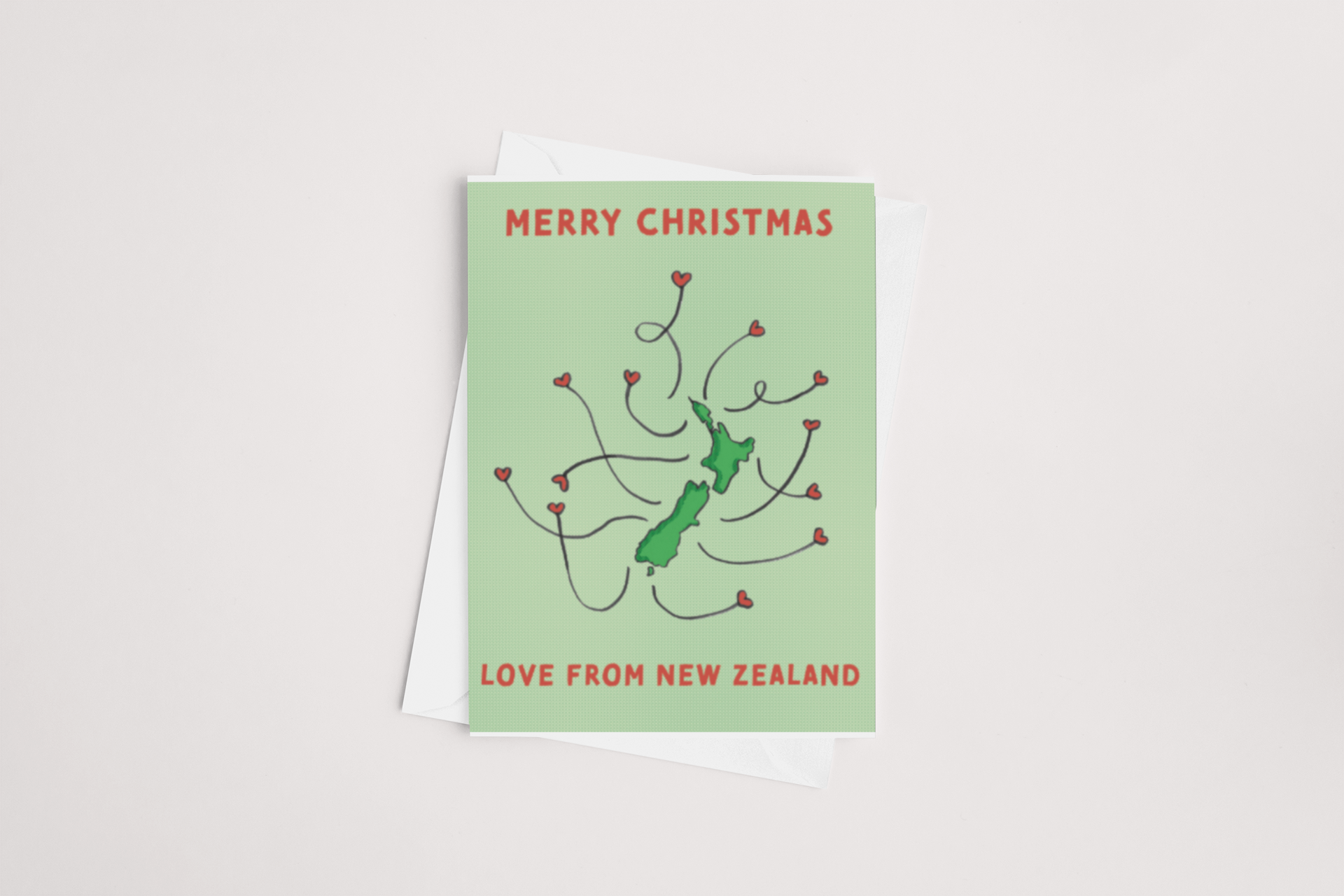 The "Love from NZ Christmas Card" by Tuesday Print features "Merry Christmas" in red above a green outline of New Zealand, with red lines and heart shapes emanating from the map. The bottom says "Love from New Zealand" in red. The card is blank inside for your personal message and comes with a white envelope, set against a light background.