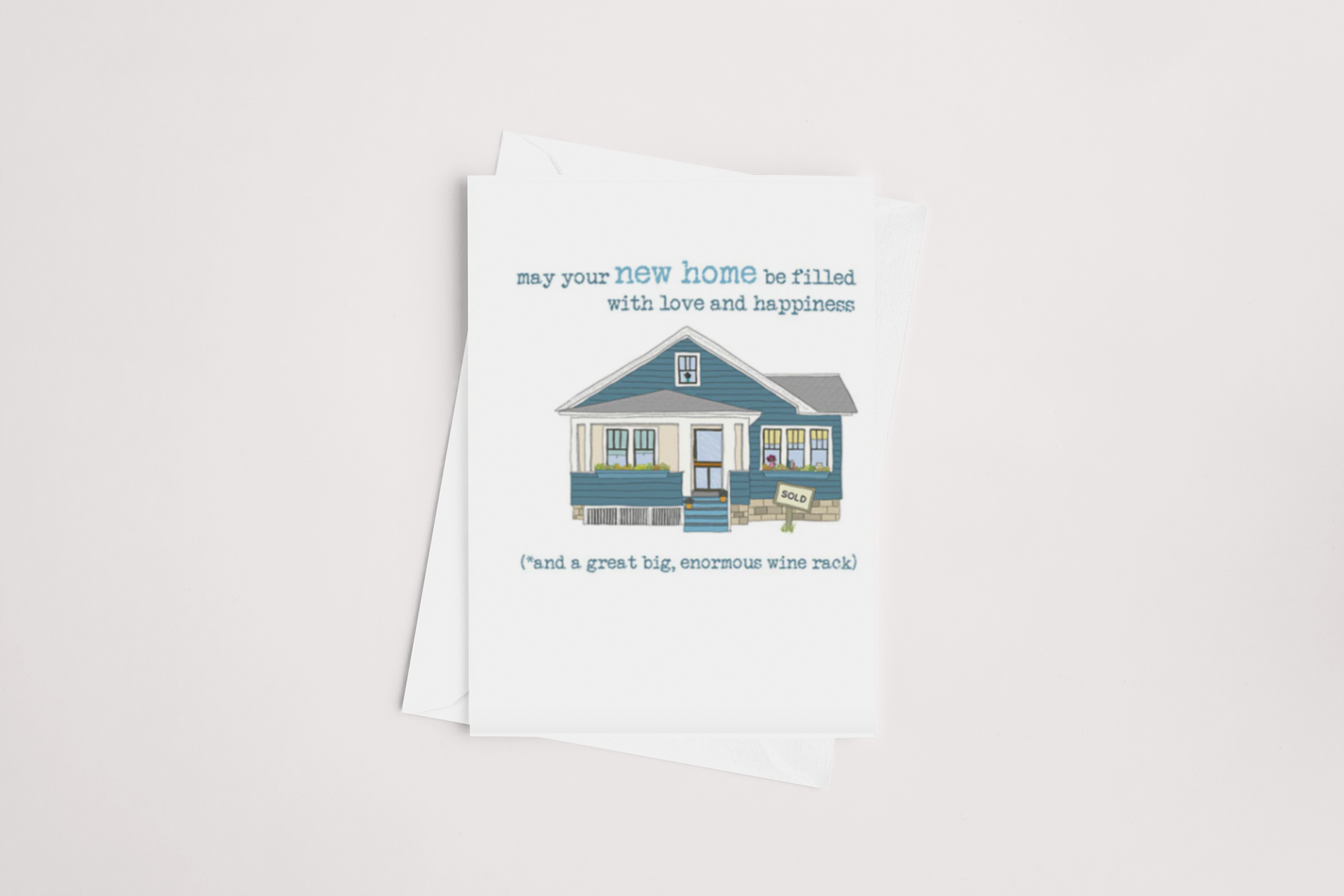 The New Home Card by Dandelion features an illustration of a blue house with a front porch, accompanied by the text: "may your new home be filled with love and happiness." Below, it continues with, "and a great big, enormous wine rack!" The card is blank inside and comes with a brown envelope against a white background.