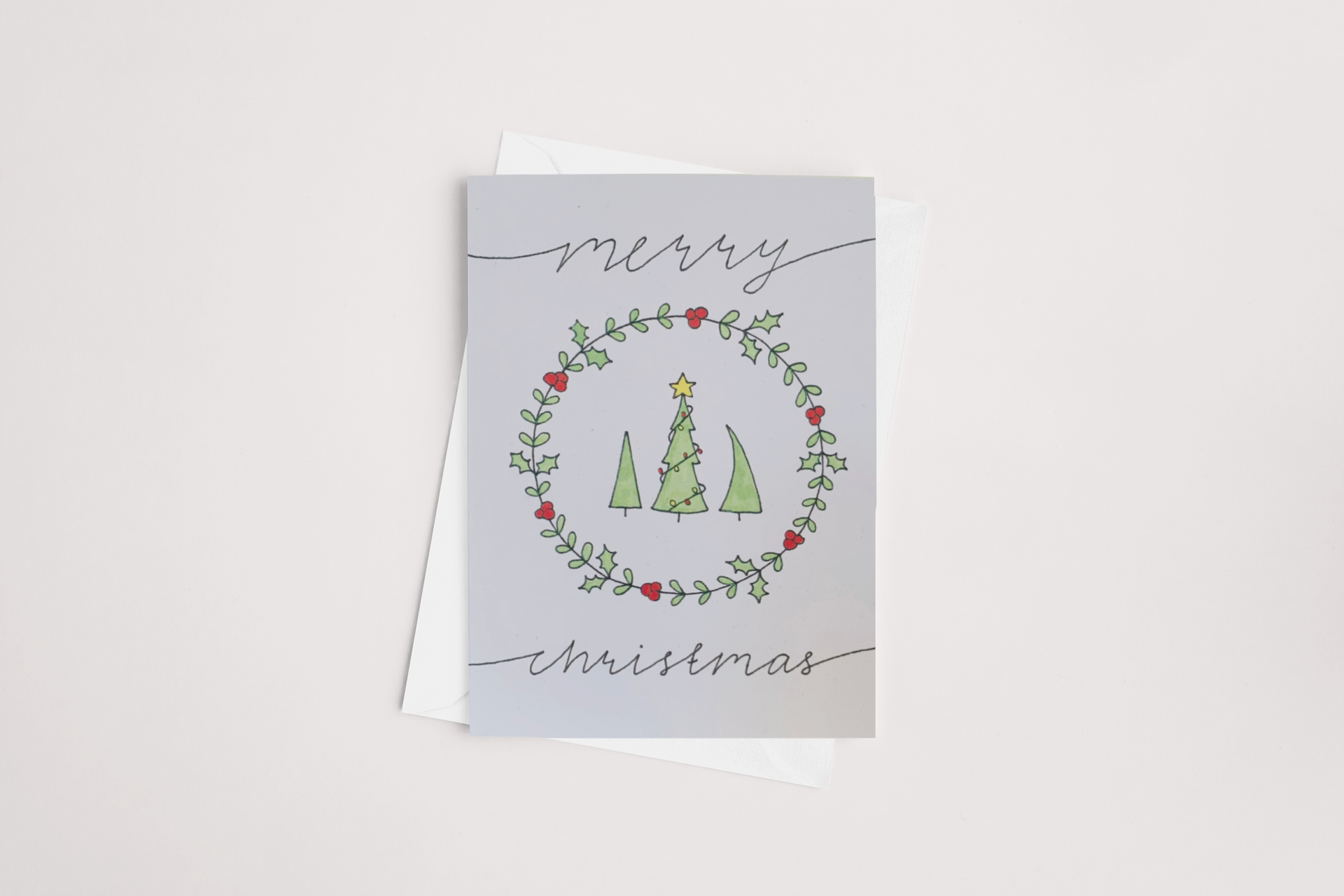 The "Merry Christmas Card" by Sweet Pea Creations features a charming wreath of green leaves and red berries encircling three green trees, each crowned with a yellow star. The words "Merry" and "Christmas" are handwritten above and beneath the wreath for an elegant touch. It comes with a white envelope, is blank inside for your personal message, and is available in a multibuy offer.