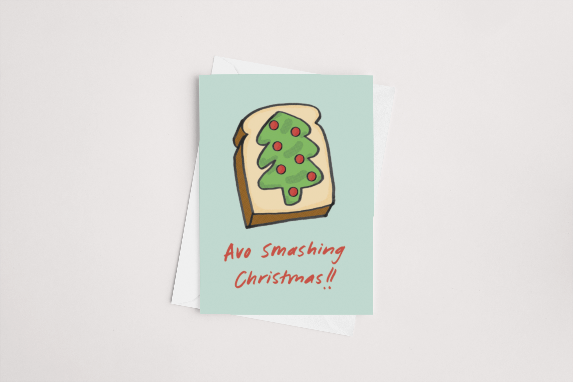 The "Avo Smashing Christmas Card" by Tuesday Print showcases a whimsical illustration of avocado toast styled as a Christmas tree, adorned with festive red dots. Set on a light blue background, the card features the red cursive message "Avo Smashing Christmas!!" and includes a partially visible envelope. The inside is left blank for you to add your personal message.