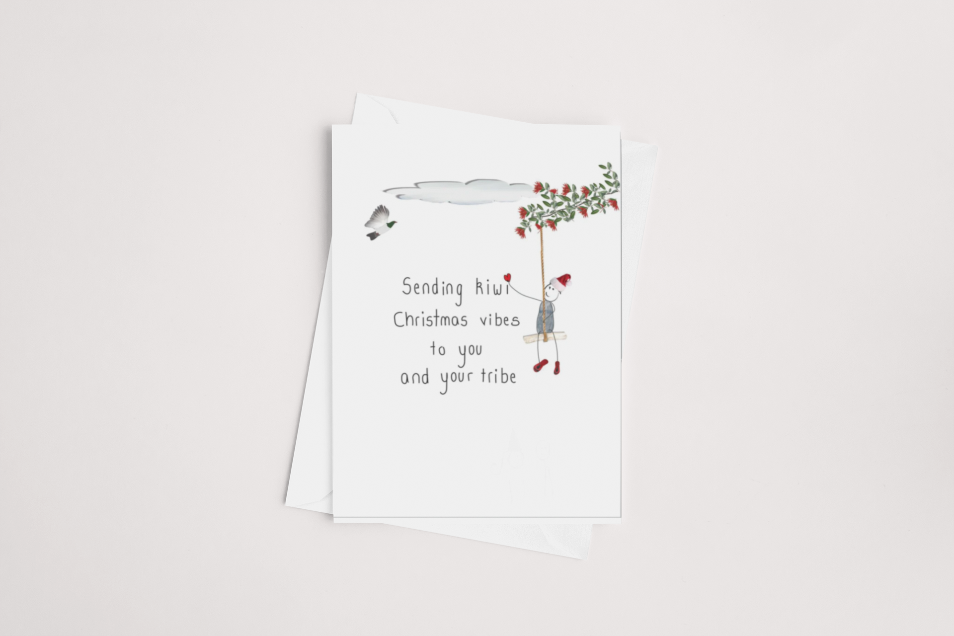 The Vibes Christmas Card by iCandy showcases a whimsical illustration of a person hanging from a sprig of holly. The greeting says, "Sending hiiiu Christmas vibes to you and your tribe." This delightful message card is displayed on a light beige surface with its accompanying white envelope tucked underneath.