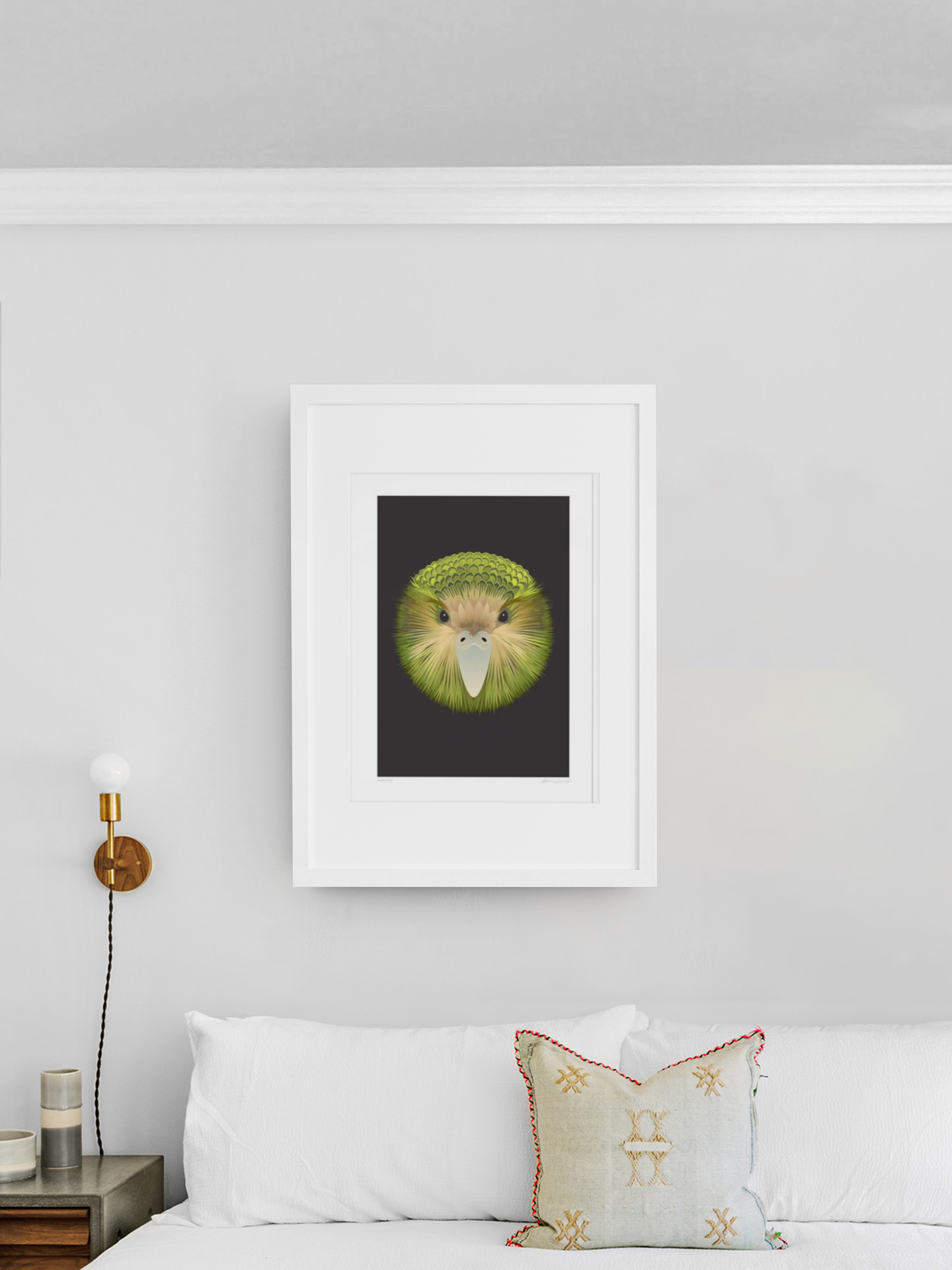 A contemporary bedroom showcases a white bed adorned with a textured pillow, positioned under a white-framed print titled "Kakapo" by Glenn Jones. The art piece intriguingly depicts a lion's face crafted from a kiwi slice. The walls are painted in light gray, and to the left is a round wall lamp along with a small table decorated with various items.