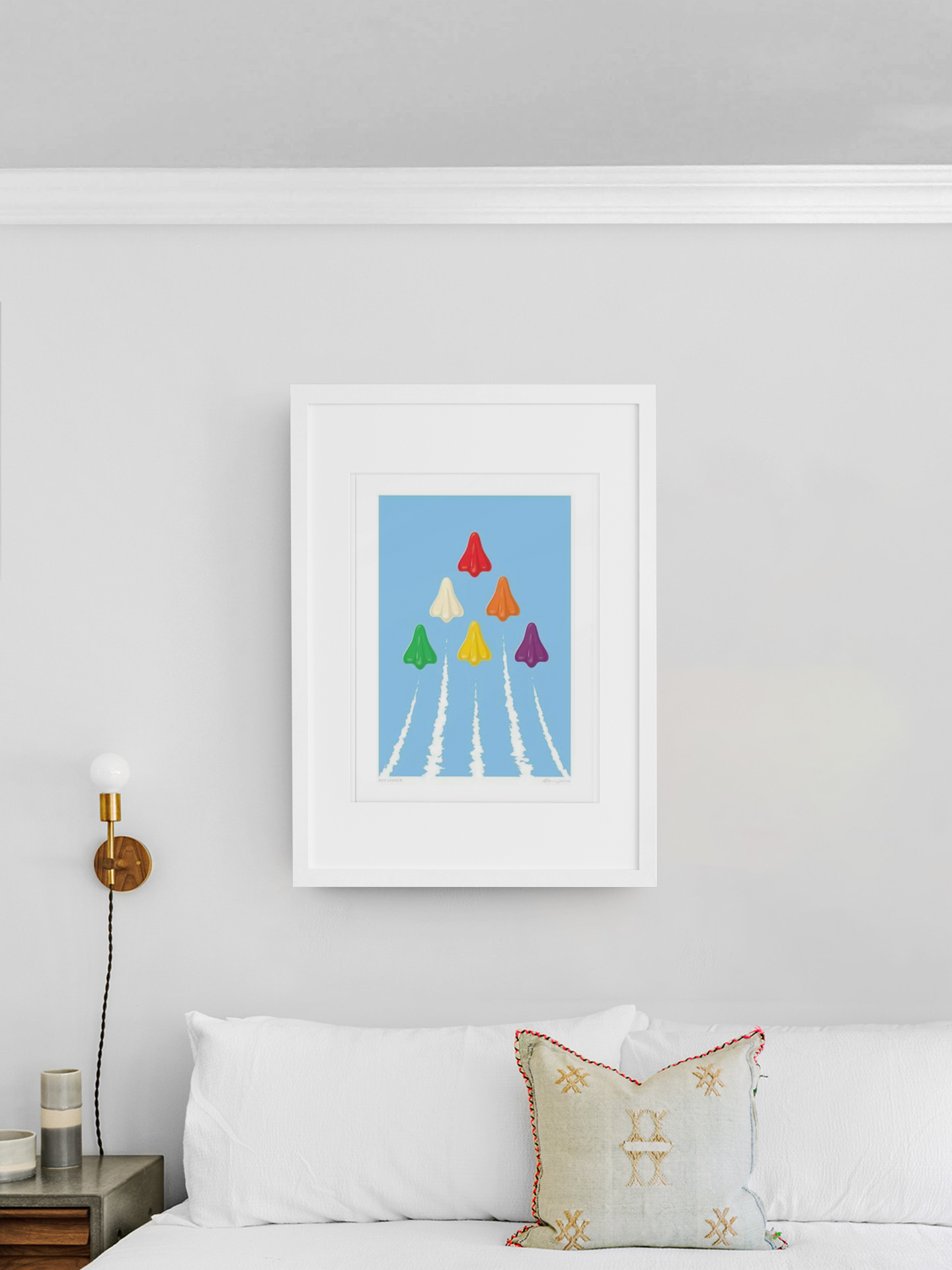 A framed print titled "Red Leader" by Glenn Jones hangs on a light gray wall above a white bed. The artwork showcases colorful paper airplanes soaring against a blue sky, each leaving a white contrail. A small wall lamp and decorative pillow adorn the bed.