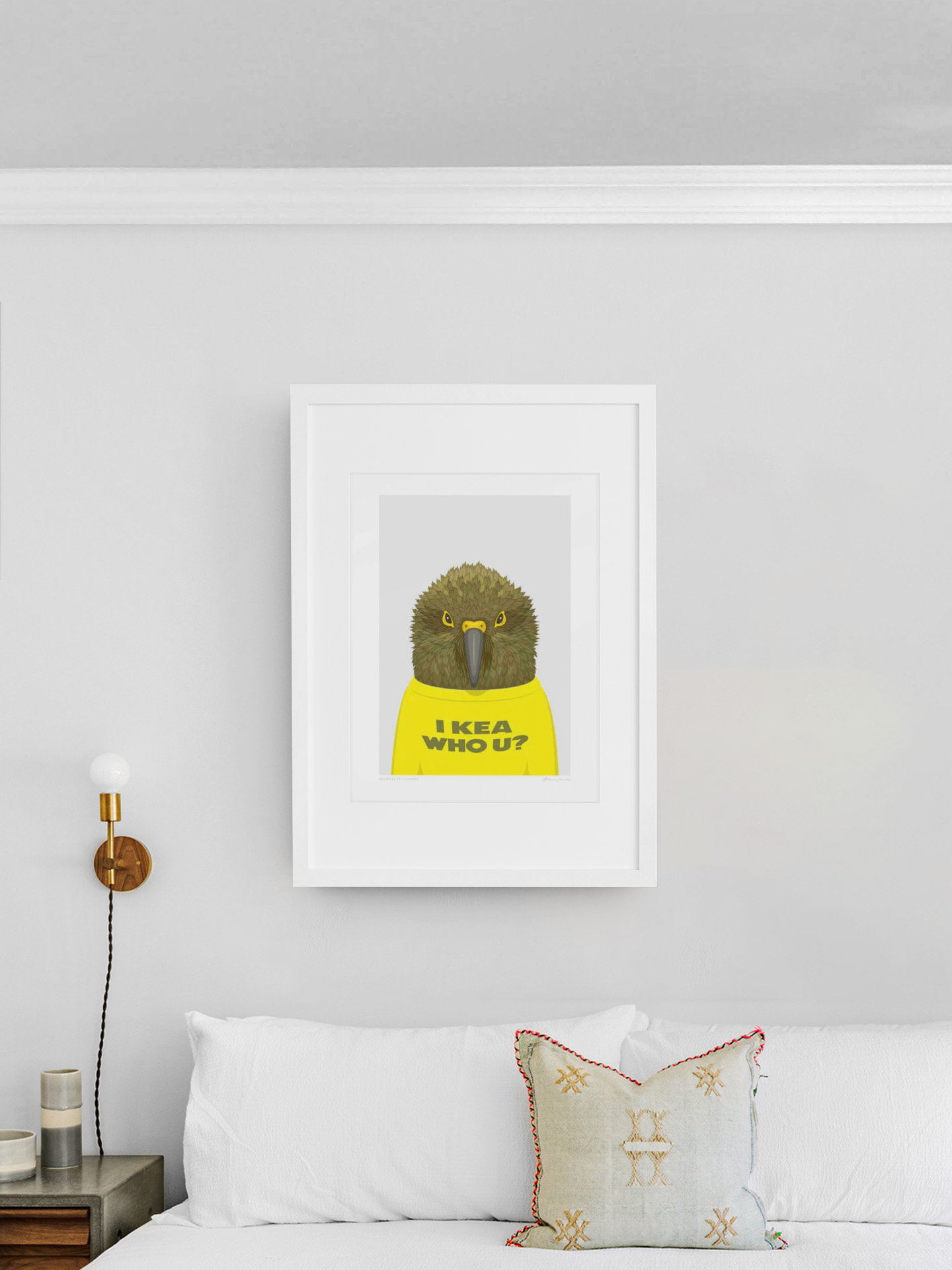 A minimalist bedroom showcases "Animals in T-Shirts - Kea by Glenn Jones" as framed artwork on the wall above a white bed adorned with a patterned cushion. The artwork portrays a baboon in a yellow shirt with the words "I KEEA WHO U?" There is also a single wall-mounted lamp featuring a brass finish on the left side.