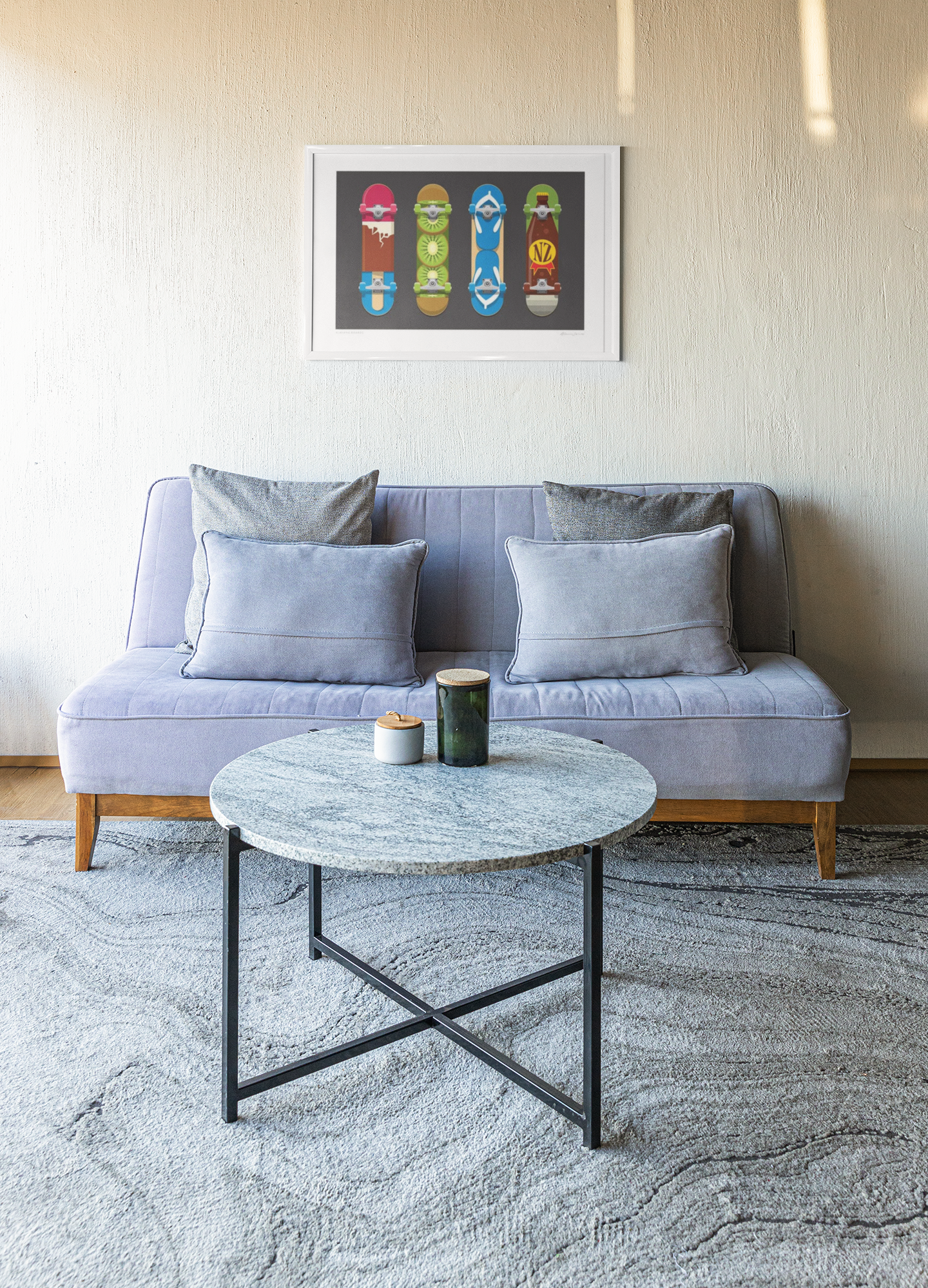 In a modern living room, there's a light gray sofa adorned with two large cushions. Hanging above the sofa is the "Kiwiana Boards" artwork by Glenn Jones, showcasing colorful skateboard decks in his distinct nostalgic style. A round marble coffee table with black metal legs is situated on a textured gray rug, holding both a black and white candle.