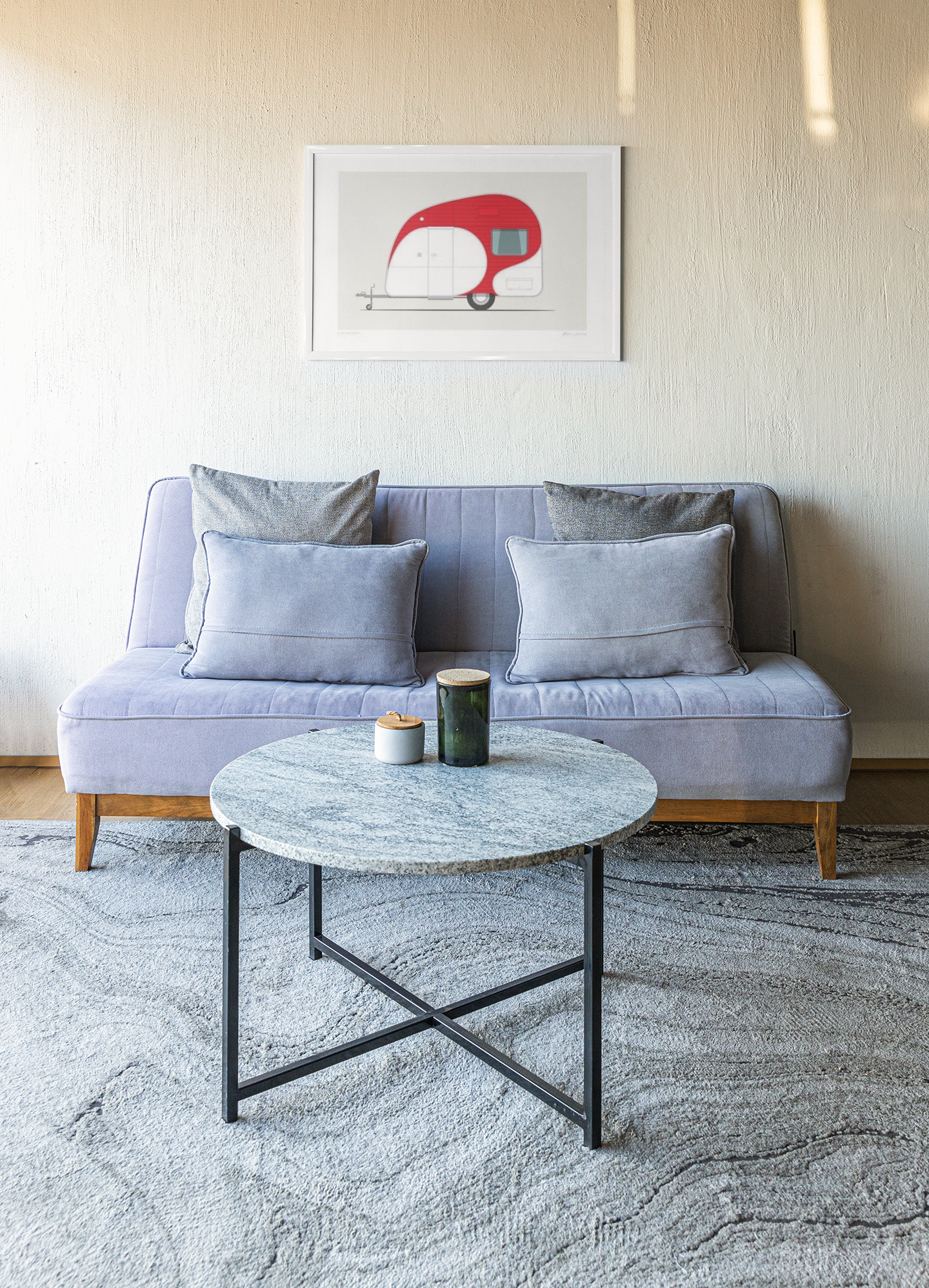 A modern living room features a light gray sofa with matching cushions. Above it, a Kiwi Getaway framed print by Glenn Jones, depicting a red camper trailer, adds charm. In front, a round marble coffee table with a black metal frame displays two candles, all set on a textured gray rug to enhance elegance.