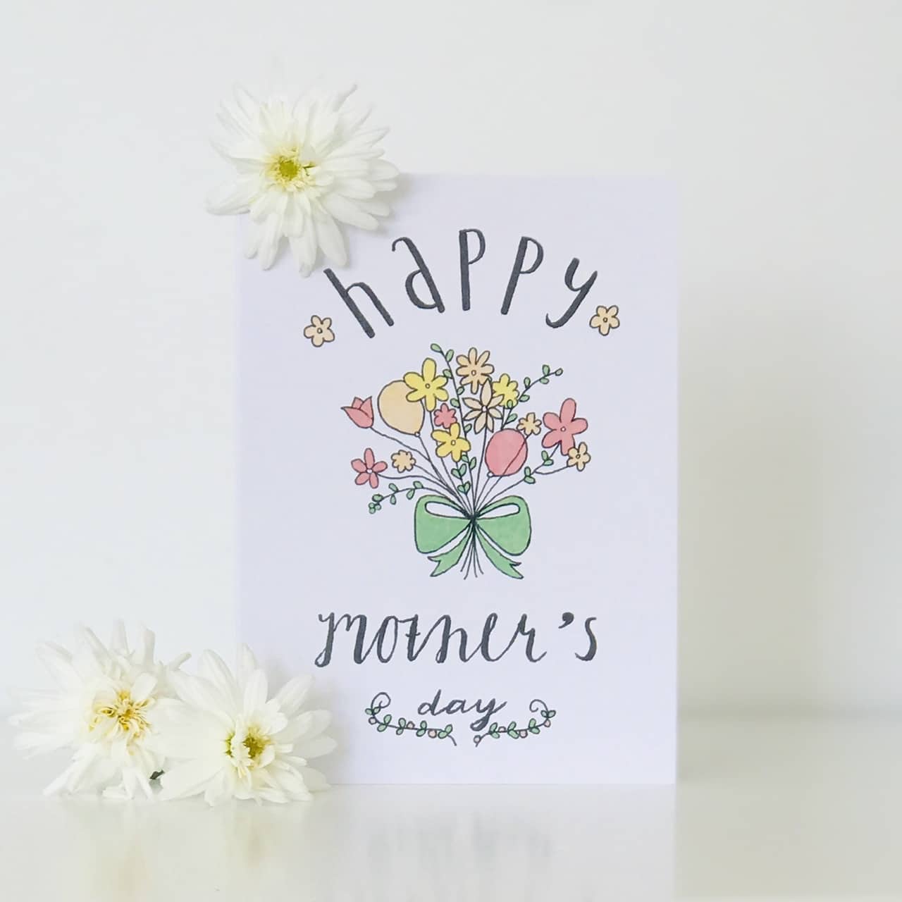 A charming Floral Mother's Day Card from Sweet Pea Creations with a floral design and calligraphy standing upright on a white surface, flanked by delicate white flowers.